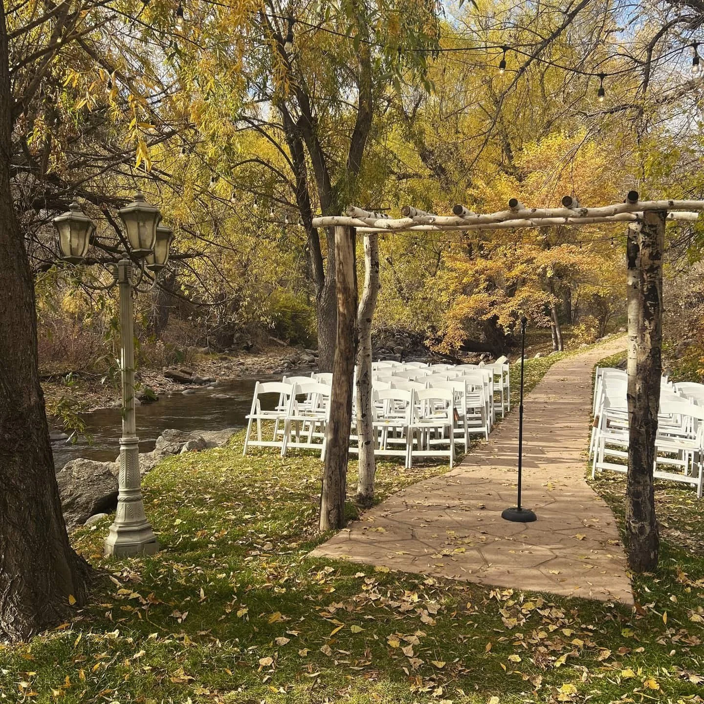 The outdoor ceremony site at Boulder Creek, situated next to the gentle creek. There are rows of white folding chairs on either side of the stone path that leads to the wooden arch at the end.