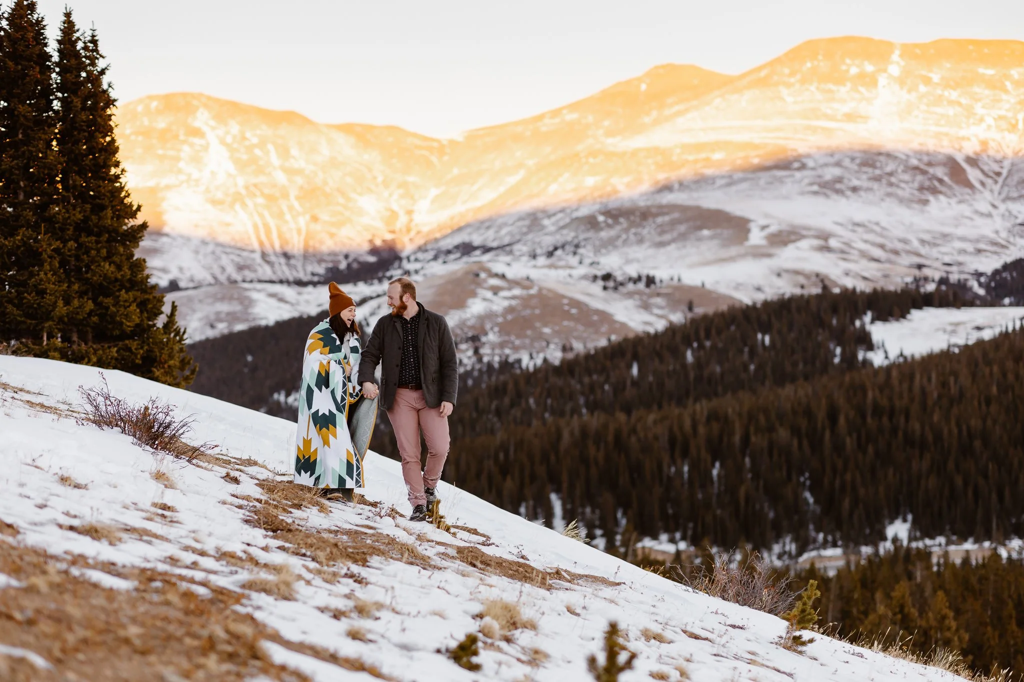 During their engagement session in the mountains of Colorado, the glowing couple hold hands while walking through the light snow that dusts the ground. The sunset illuminates the mountains behind them in golden light as the two share their excitement