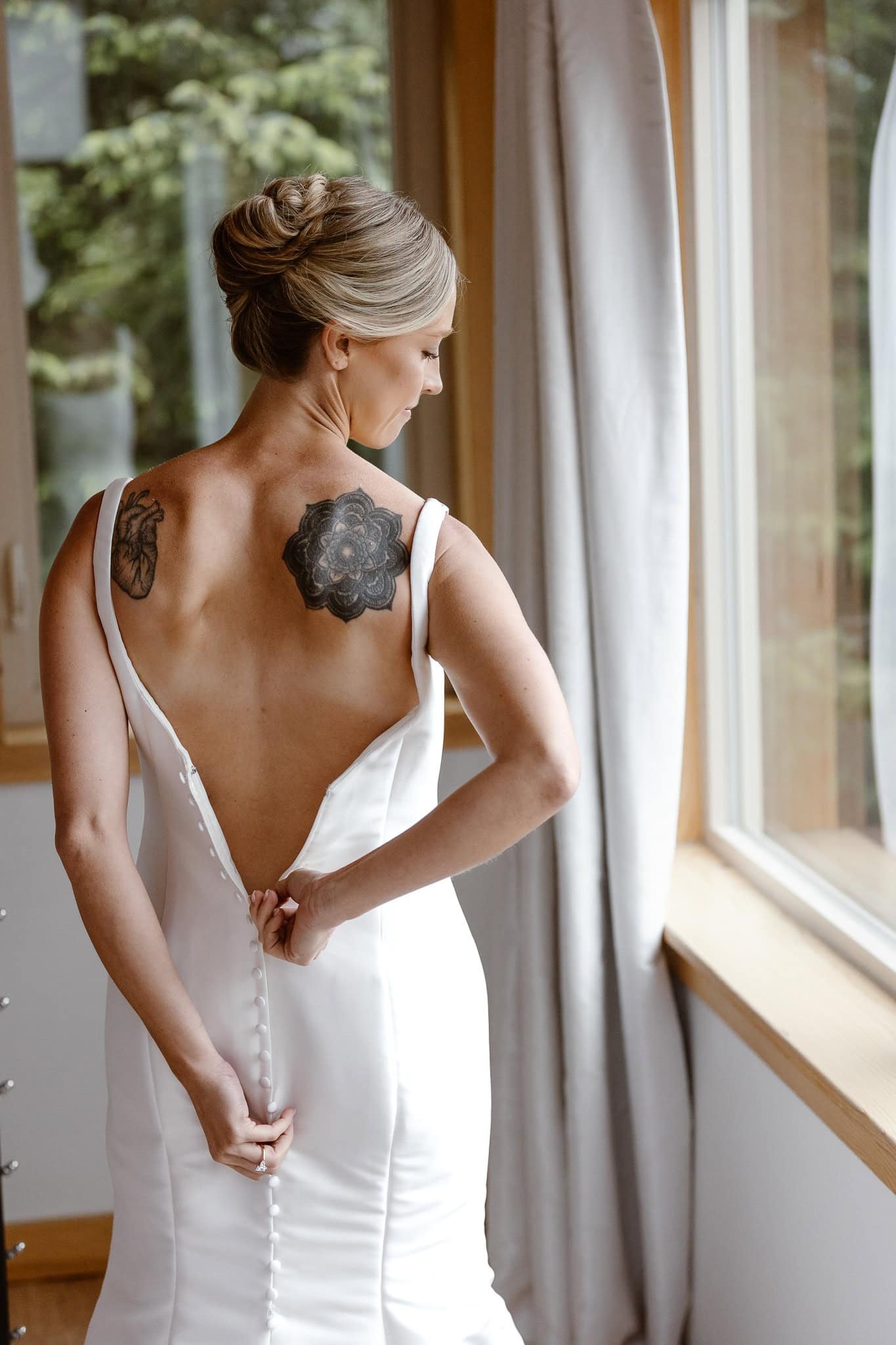 With her hair and makeup finished, the bride is seen from behind as the zips up the back of her open-back, satin, mermaid wedding dress, complete with buttons along the zipper. She has two tattoos on either side of her upper back, perfectly in view.