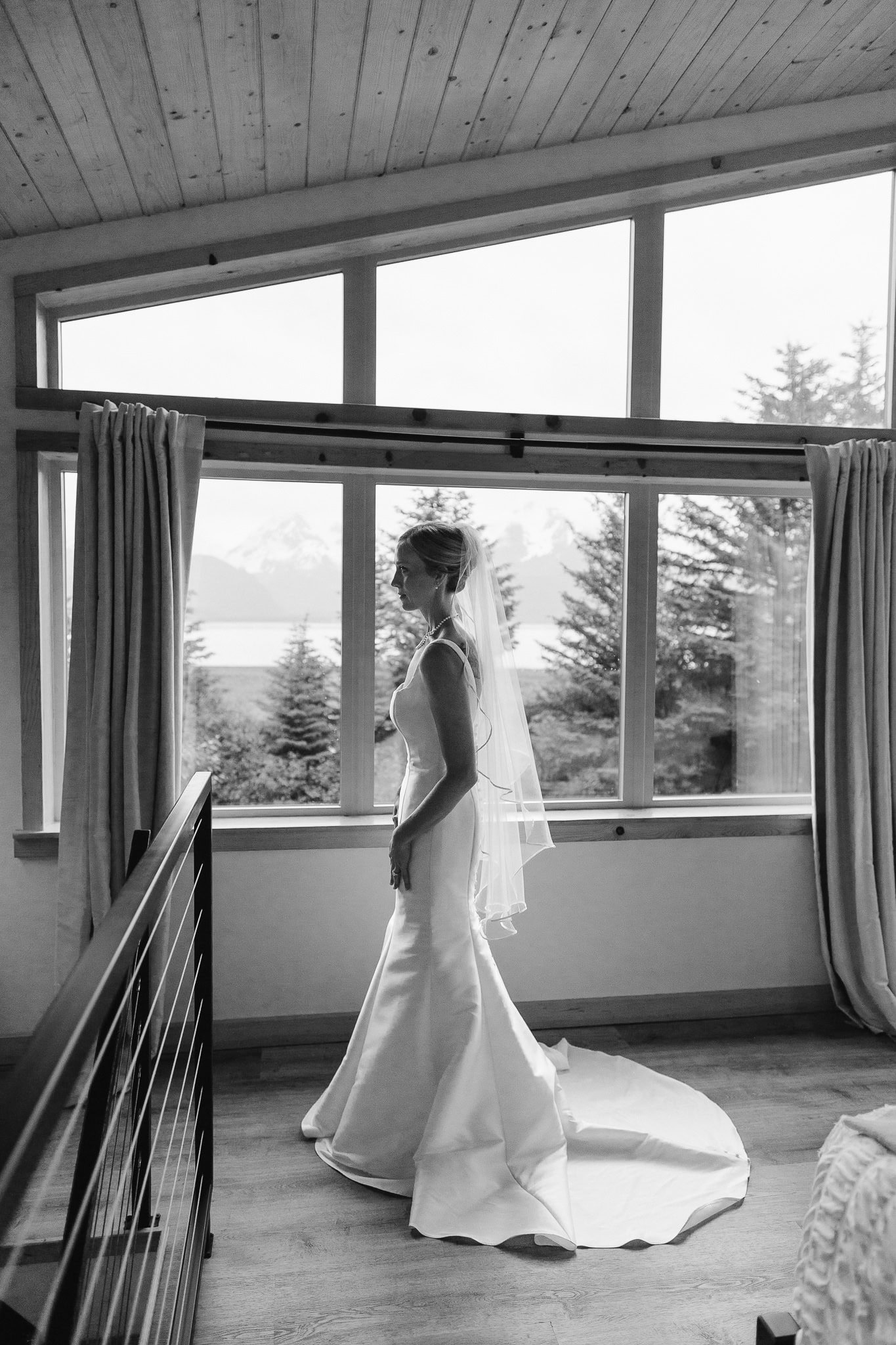 Ready for the first look with her fiance, the gorgeous bride stands in her dress, pearls, and veil, in front of a wall of windows, where you can see the snowy mountains and Resurrection Bay in the background.
