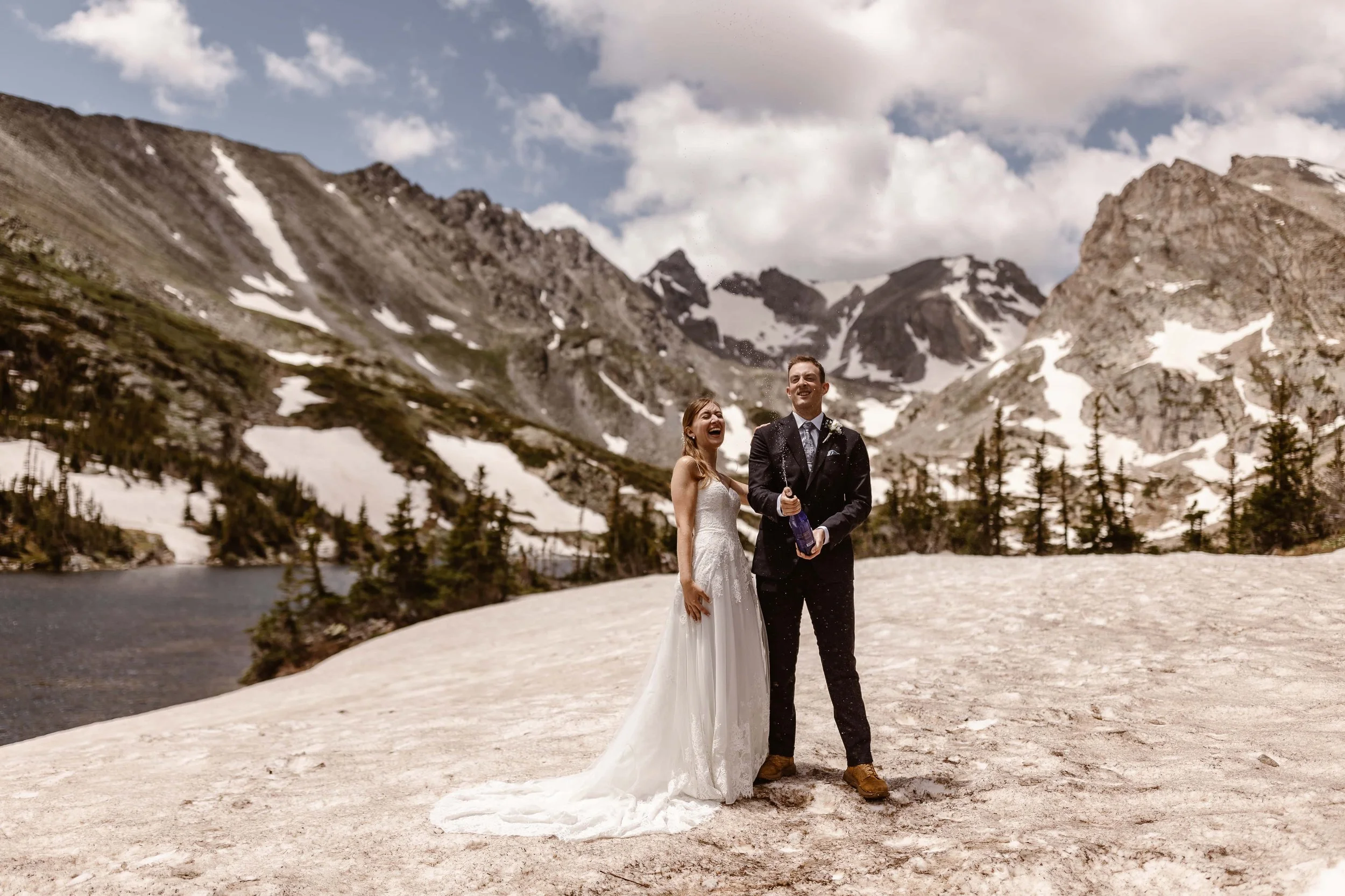 Celebrating their nuptials, the groom sprays champagne in the air while his wife stands next to him, laughing joyfully as he enjoys this moment. The gray mountains of Lake Isabelle fill the background and a beautiful summer afternoon in Colorado.