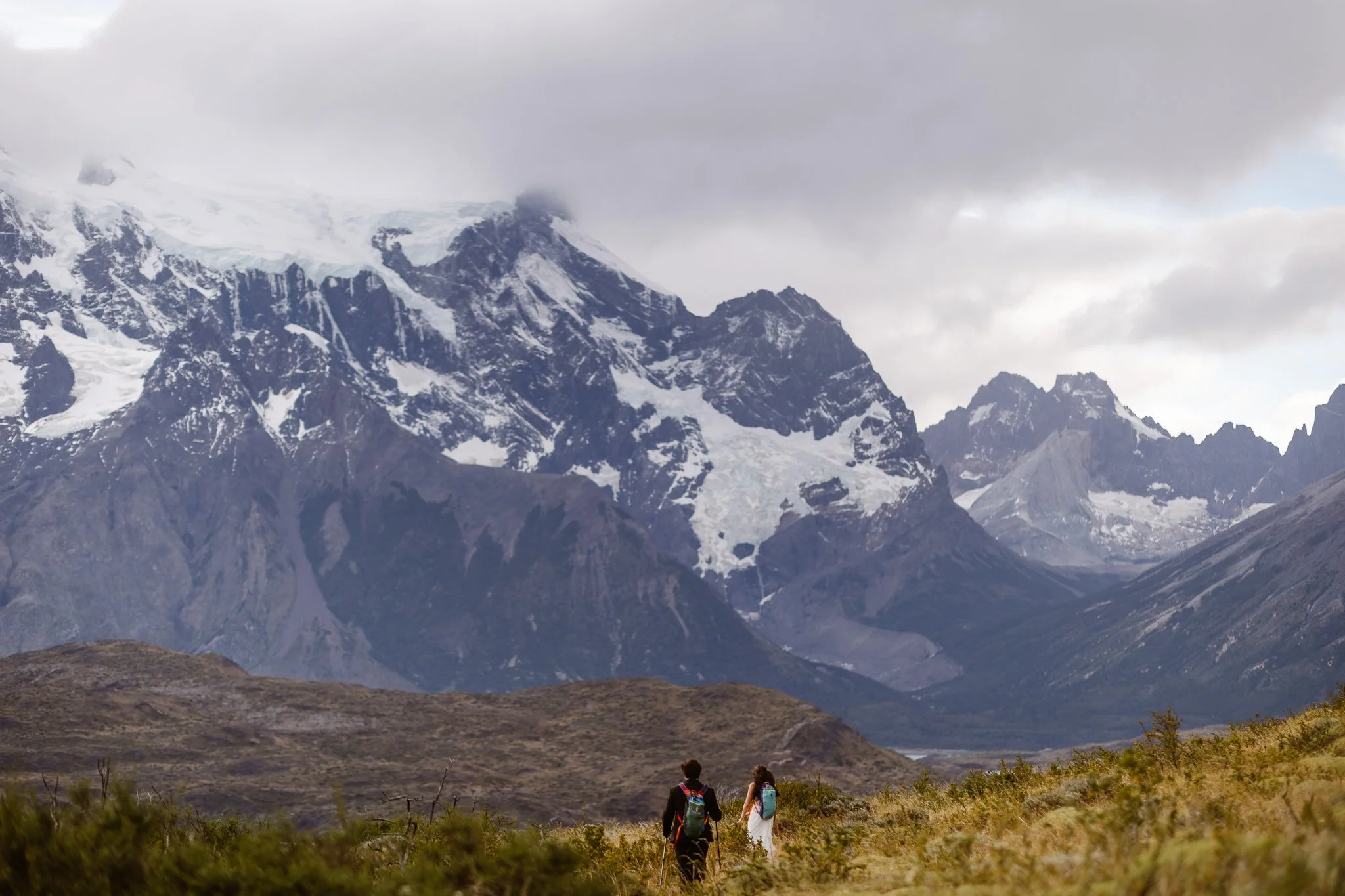 A man and woman with backpacks hike across a field toward snow-covered mountain peaks in Patagonia, Chile