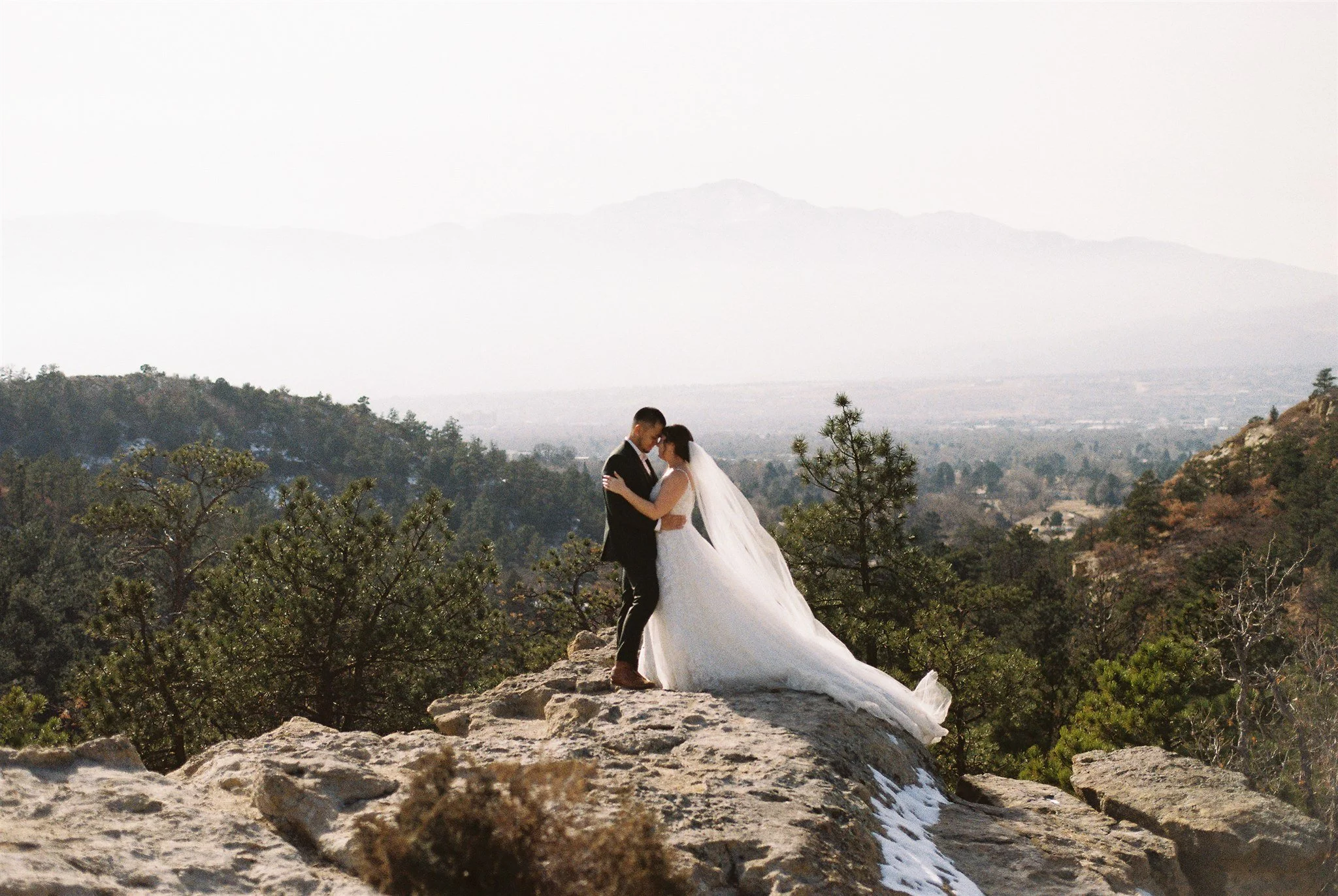 Standing closely on a rocky cliff in Colorado Springs, this bride and groom hold each other tightly during their winter elopement. Behind them is a sea of green trees and the barely visible mountains in the distance.