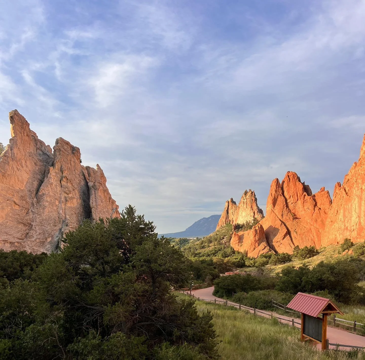 A view of the towering orange rock formations at the Garden of the Gods. Between the two formations is a flat red walking path for visitors, with lush trees, bushes, and grass filling the space around it.