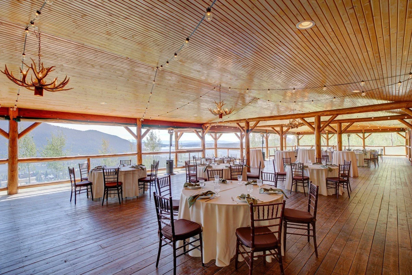 The reception space of Grand Lake Lodge with floor to ceiling wood paneling, antler chandeliers, and round tables with wooden chairs. The sides are open to the elements and show off the gorgeous view from this pavilion.