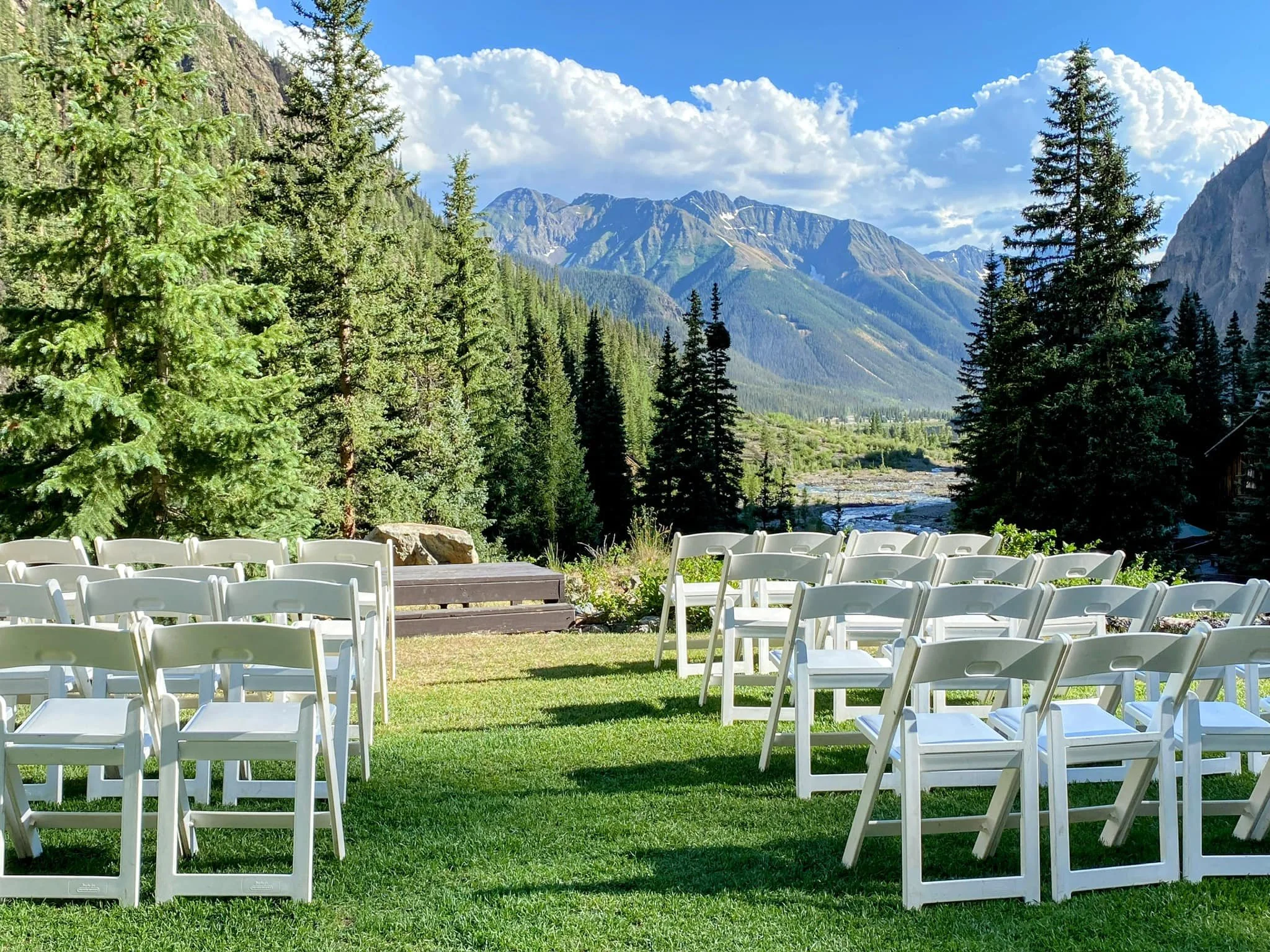 Rows of white folding chairs are aligned on the grass before a raised wooden platform for wedding ceremonies, with the view of the San Juan mountains and gentle creek before them.