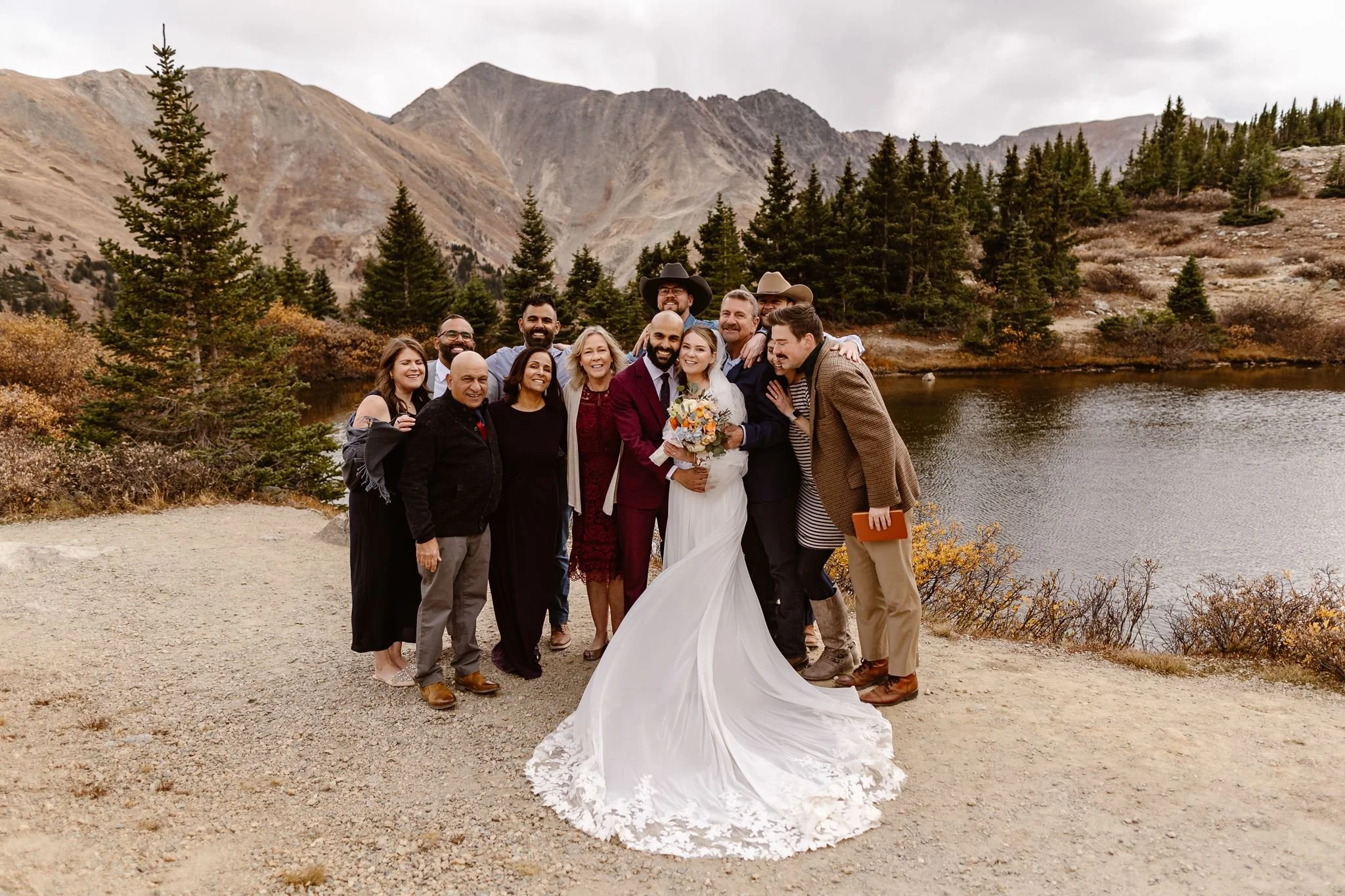 In celebration of their unity, the bride and groom hug each other tightly as their guests surround them closely for a group photo. The bride holds her orange, blue, ivory, and green bouquet that pops against the white of her dress.