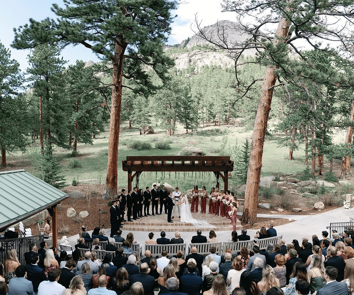 A raised stone stage off a paved path that houses a wedding ceremony, complete with a flower-adorned wooden archway. There are rows of benches full of wedding guests watching this afternoon wedding, with the tall mountains in the background.