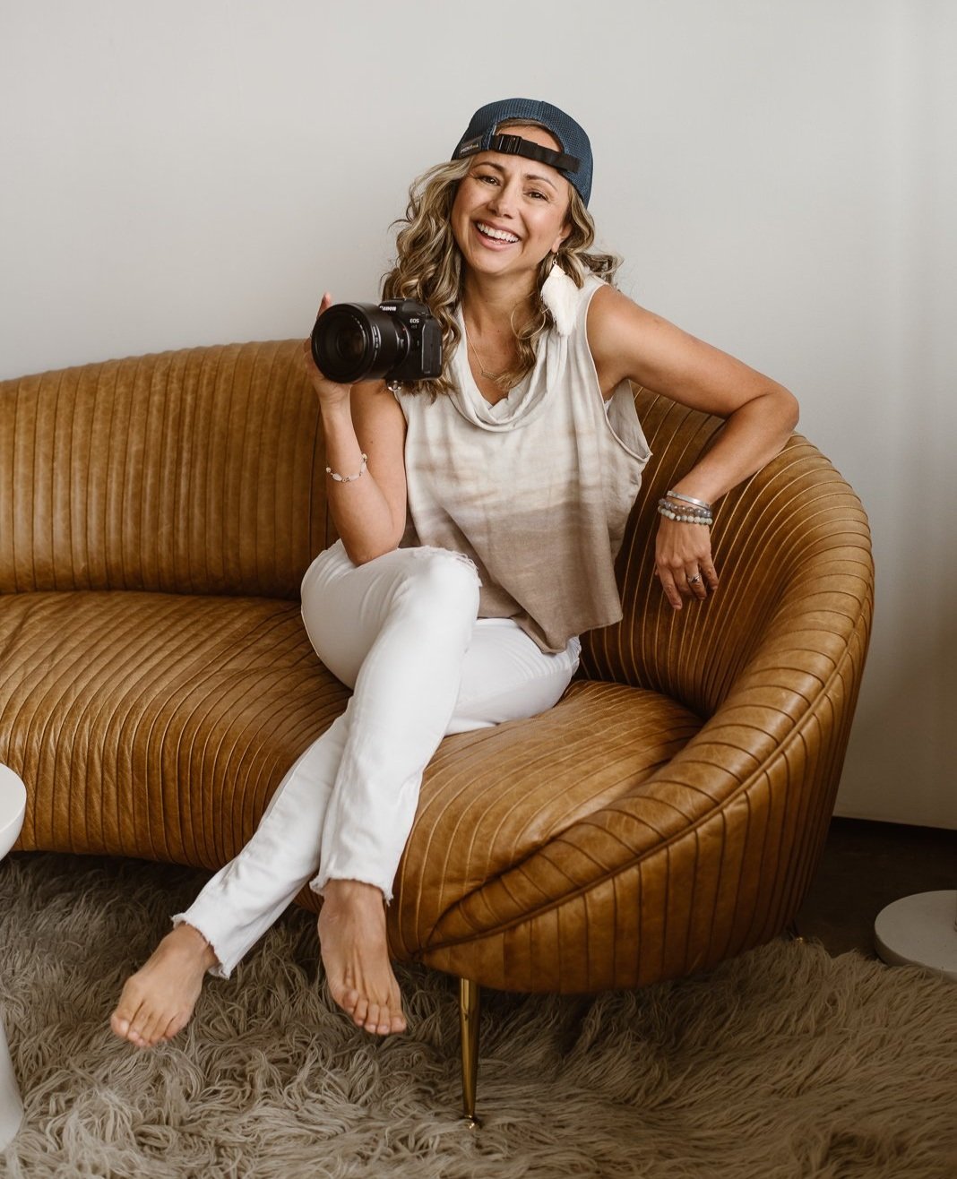 Sitting on a crescent-shaped brown leather couch, photographer Andrea Enger smiles happily while holding her camera up in right hand. She's wearing white pants and a cream/taupe loose shirt, and backwards hat.