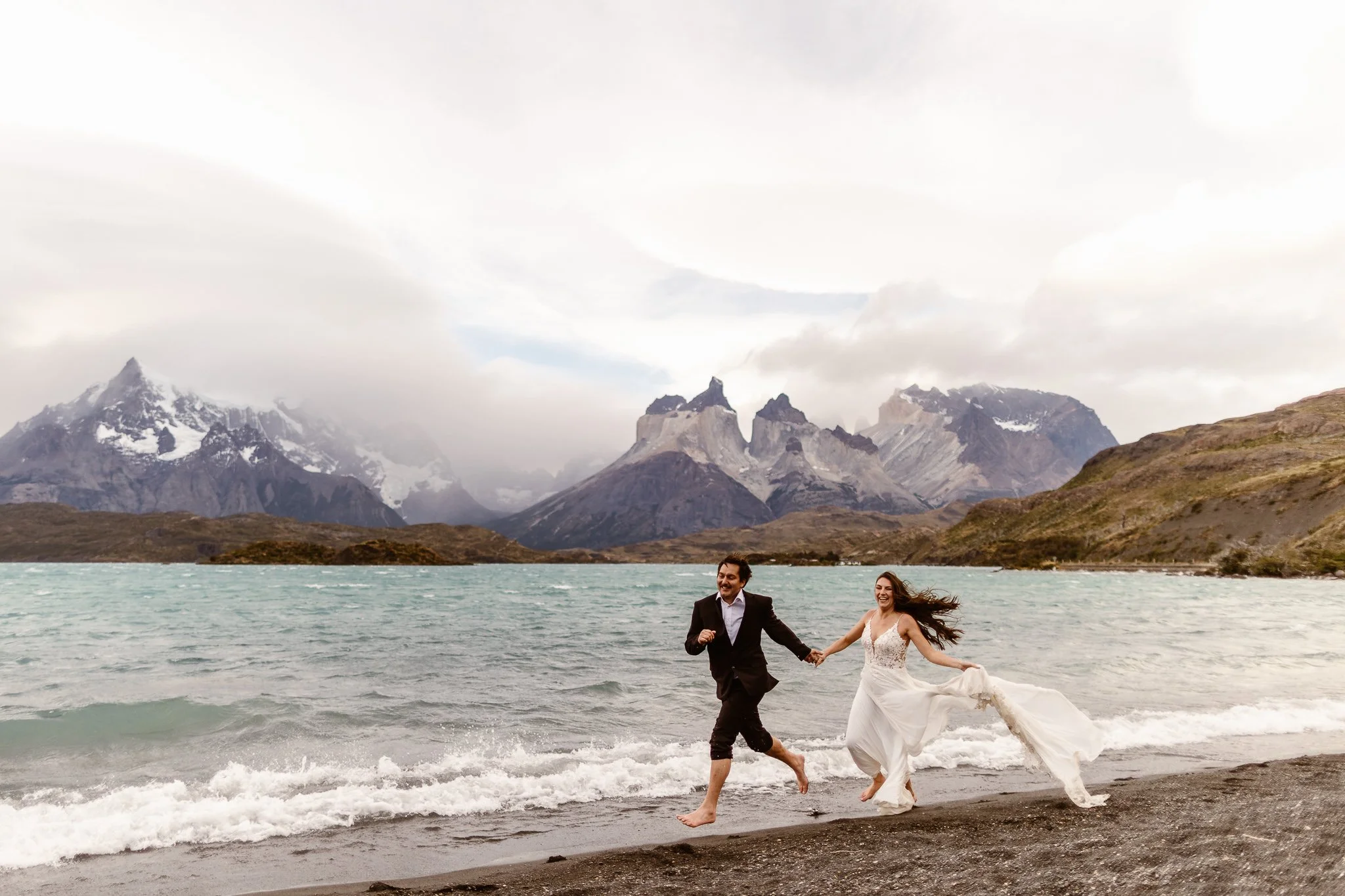 Smiling newlyweds run barefoot along the edge of a lake after their summer Patagonia elopement
