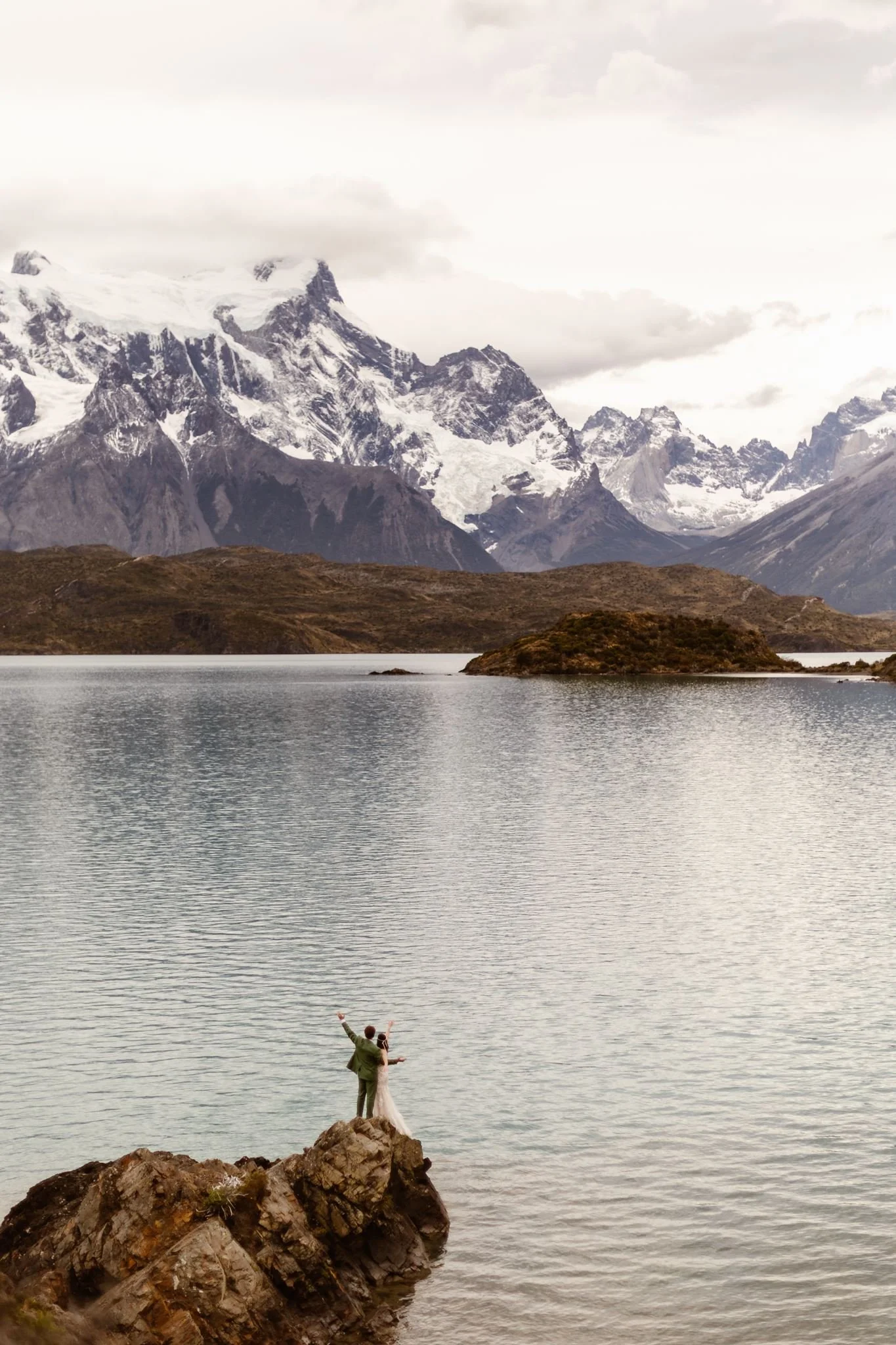 While standing on the edge of rocks on the water, the bride and groom are seen from above as they hold their arms up in the air looking away towards the mountains in the background. Here, the celebrate and thank Patagonia for the beautiful day.