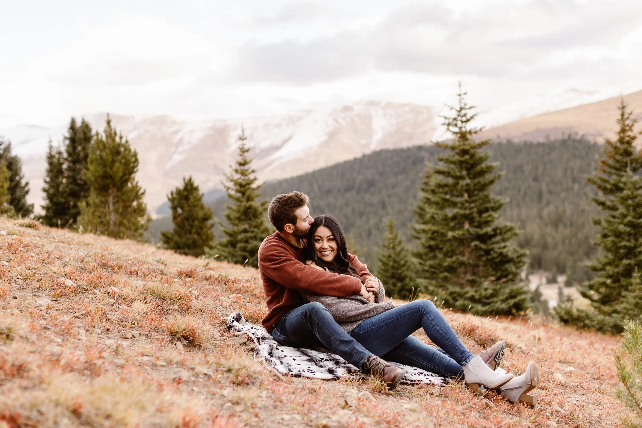 A romantic engagement session in Breckenridge amongst the fall colors of the mountains. While sitting on a picnic blanket, she leans on him as he holds her from behind, kissing her head, as she smiles brightly at the camera.