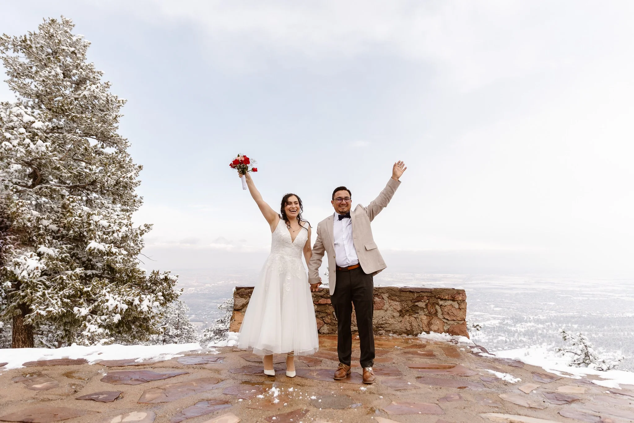 A happy bride and groom hold hands while throwing the other hand in the air, the bride holding her red and pink bouquet. They're standing in front of the small brick wall at Sunrise Amphitheater, as snow fills the background and dusts the trees.