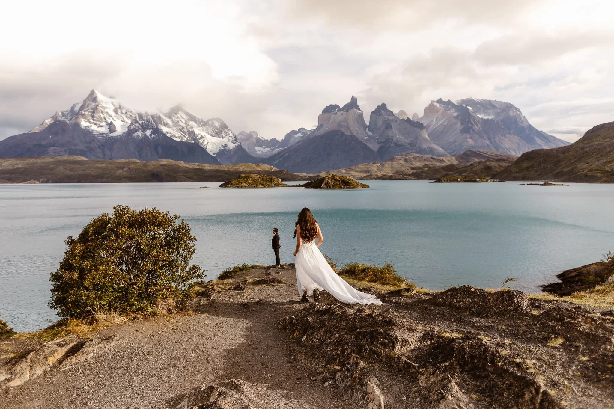 A bride walks toward her groom who stands at the edge of a lake with islands and snowy mountain peaks in Patagonia for their elopement