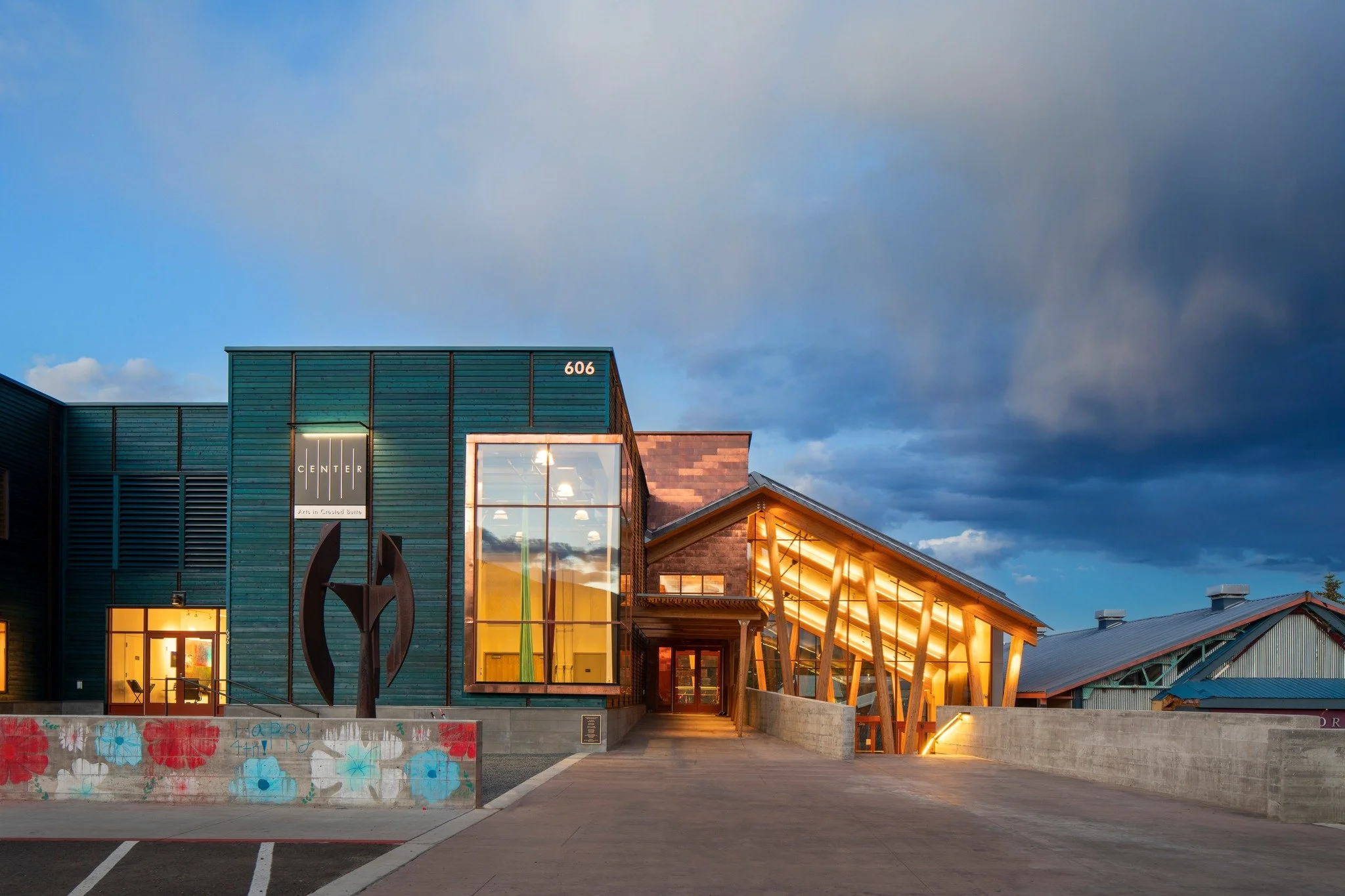 The exterior of the Center for the Arts Crested Butte during blue hour, as the building's lights glow in yellow and orange, created a warm and welcoming vibe against the teal material of the main building.