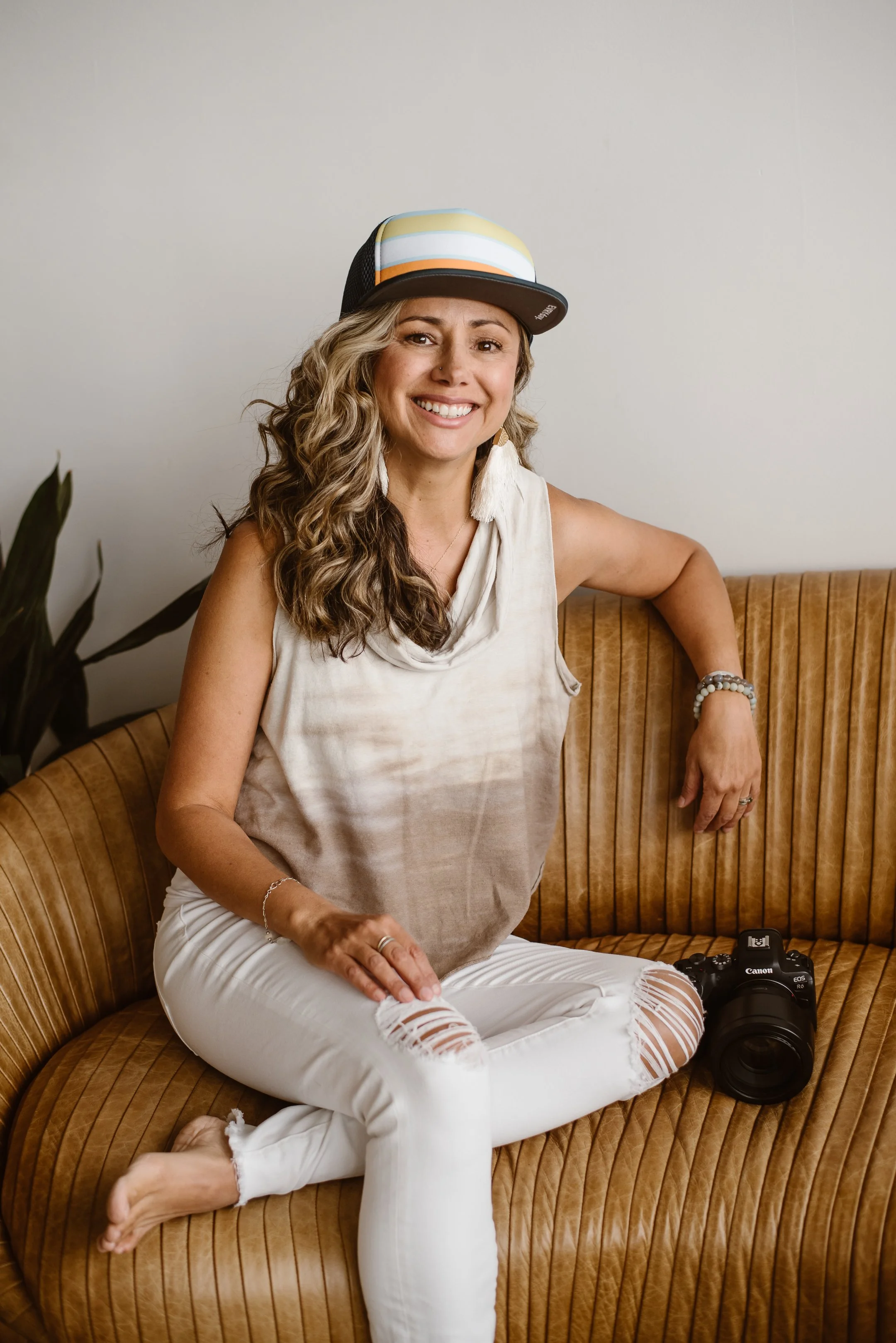 Elopement photographer, Andrea Enger, sitting on a leather couch with her left arm propped up on the back, while wearing a cream-colored shirt, pants, and striped hat, sporting a wild smile as she sits next to her camera.