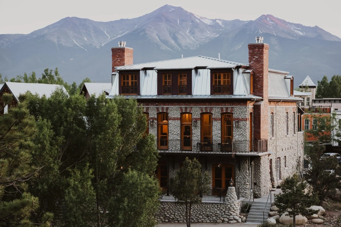 The exterior of the Surf Hotel during blue hour as the soft light kisses the stone walls and brick chimneys, along with the reddish glow on the mountain peaks in the background.