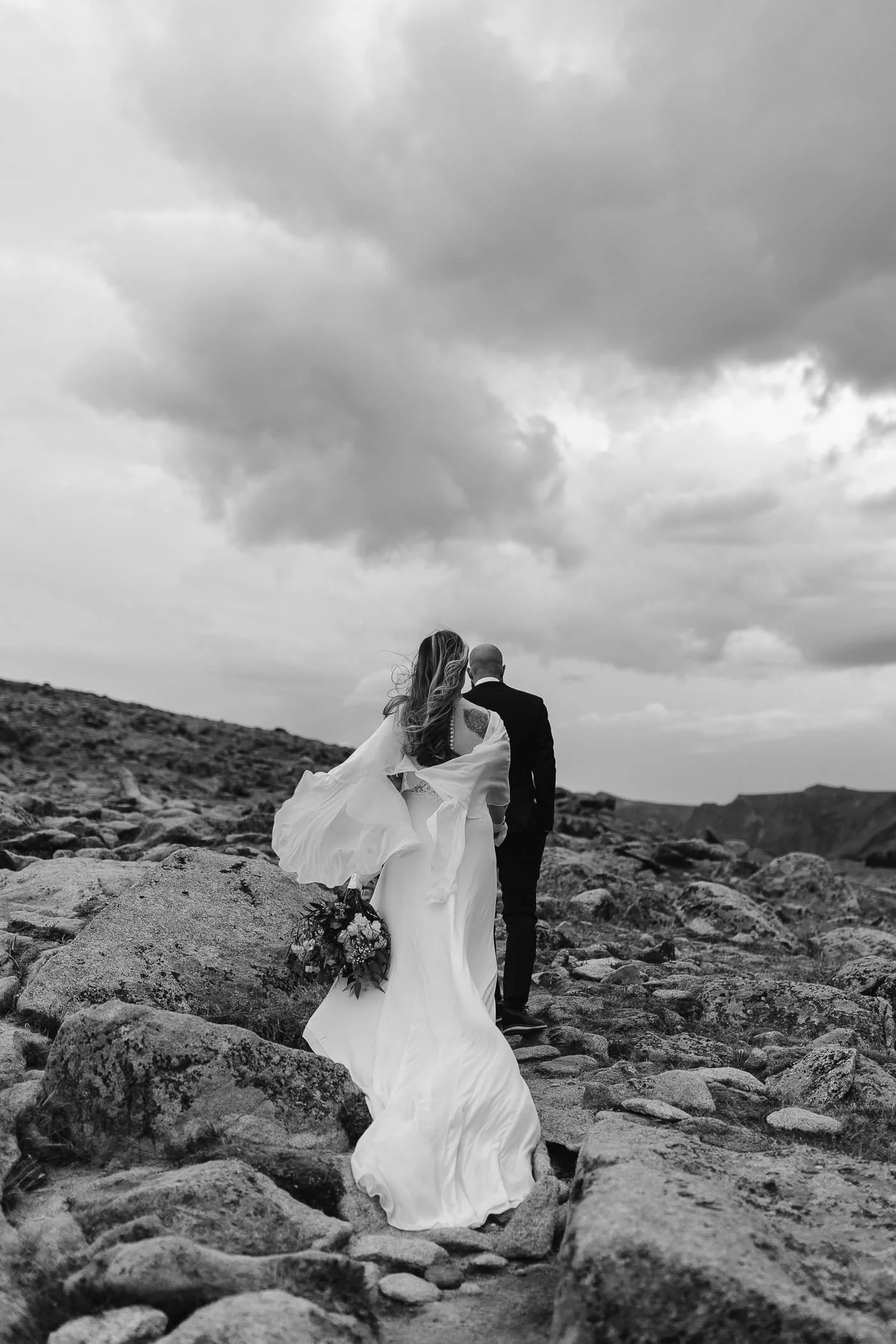 As they hike up the rocky path, the groom leads his bride as she holds his hand and follows behind. Her dress is blowing beautifully in the wind and her bouquet pops against the white of her dress.