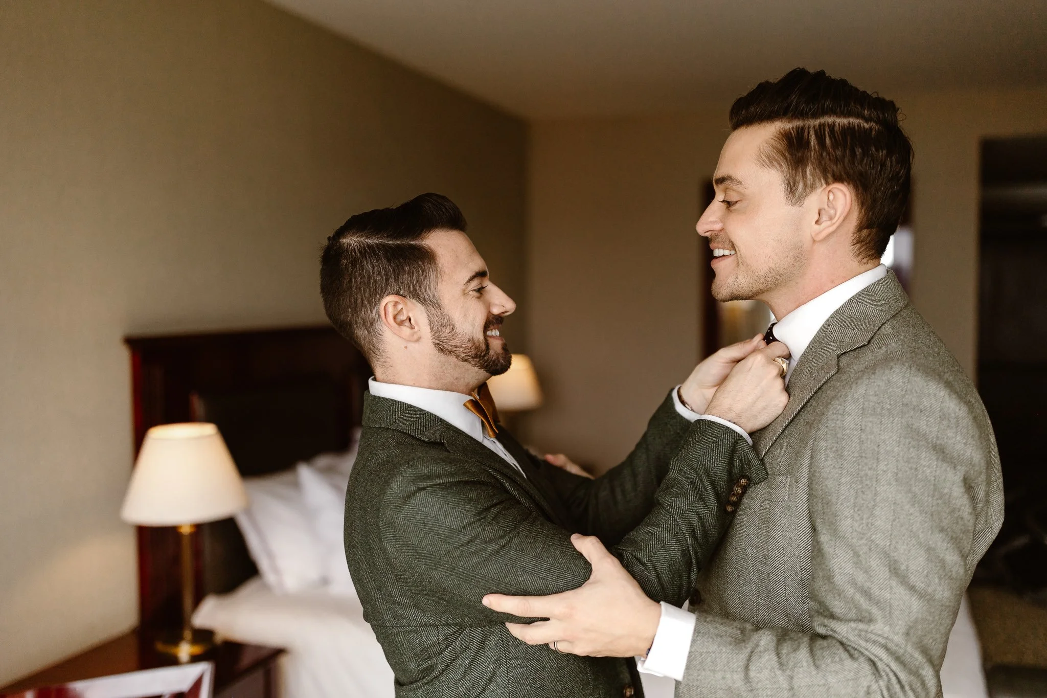 Two grooms preparing for their adventure elopement in their hotel room in Bariloche. Both wearing green suits and smiling happily as one of them adjust the tie of the other.
