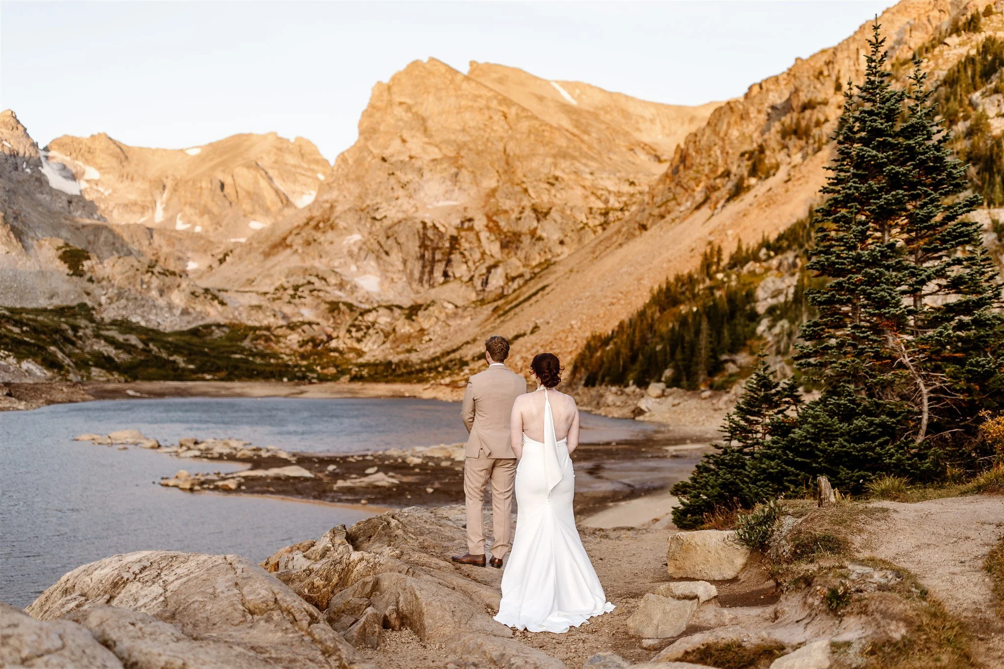 During their first look at sunrise on Lake Isabelle, surrounded by jagged peaks and rocks, the bride slowly walks up behind her groom, in a tan suit, as he stands towards the lake, looking at the mountains.