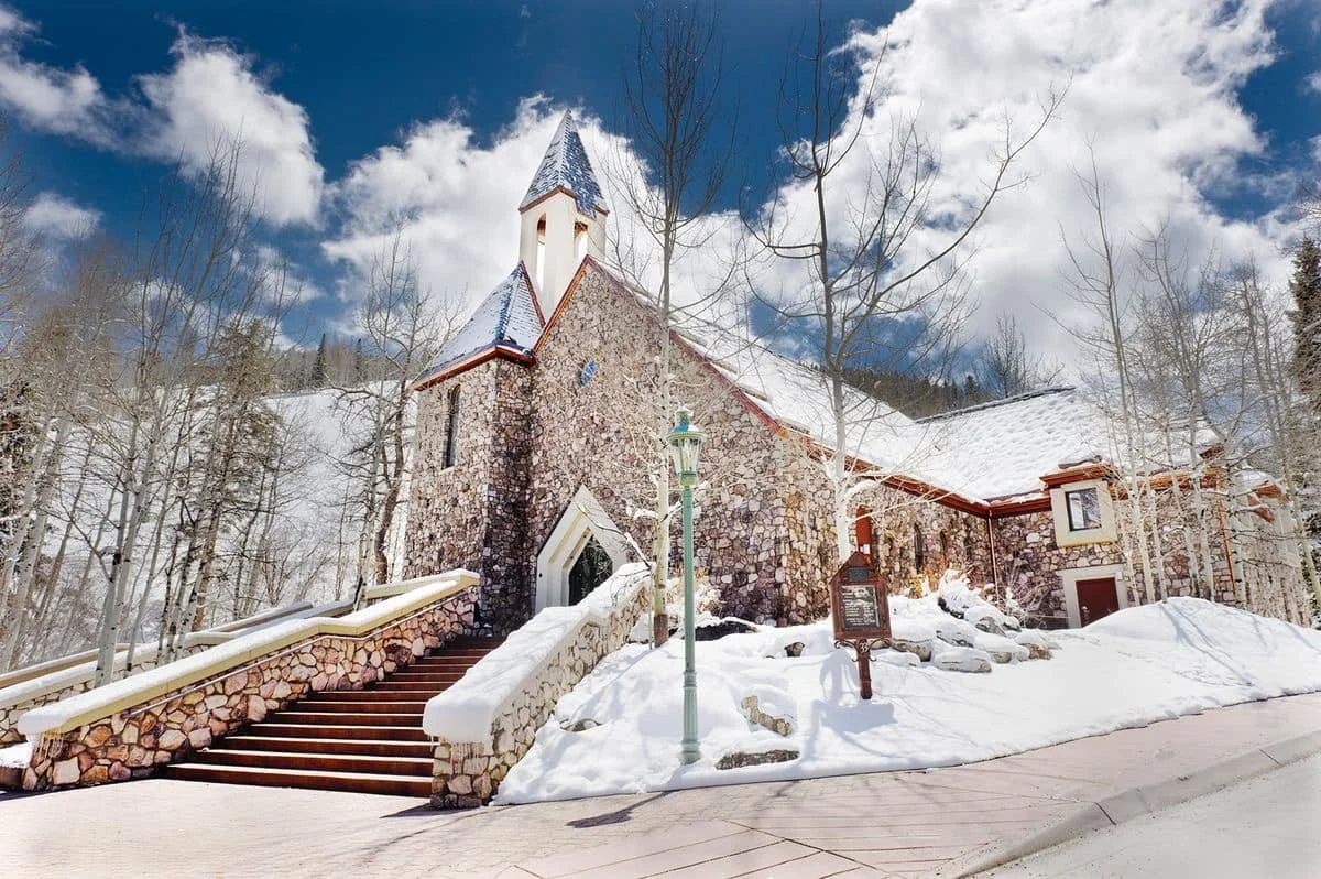The street view of the Chapel at Beaver Creek, which is a stone building with a bell tower in the front and a large staircase that leads up to the doors.
