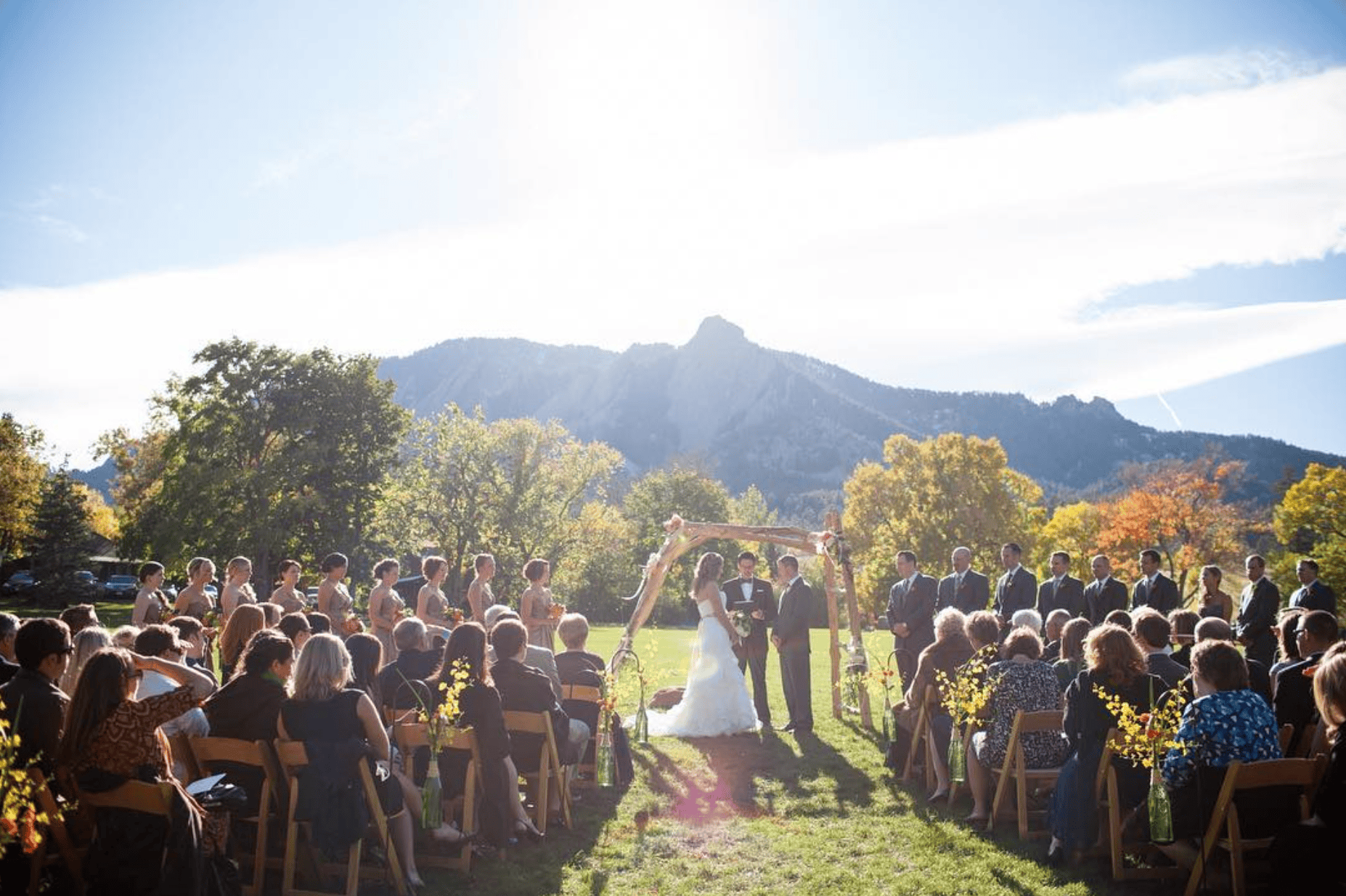 On the sprawling lawn of Rembrandt Yard, a wedding ceremony takes place under a wooden arch by the trees and towering mountains ahead. Guests are seated on either side in folding chairs and the large bridal party stand on either side of the couple.