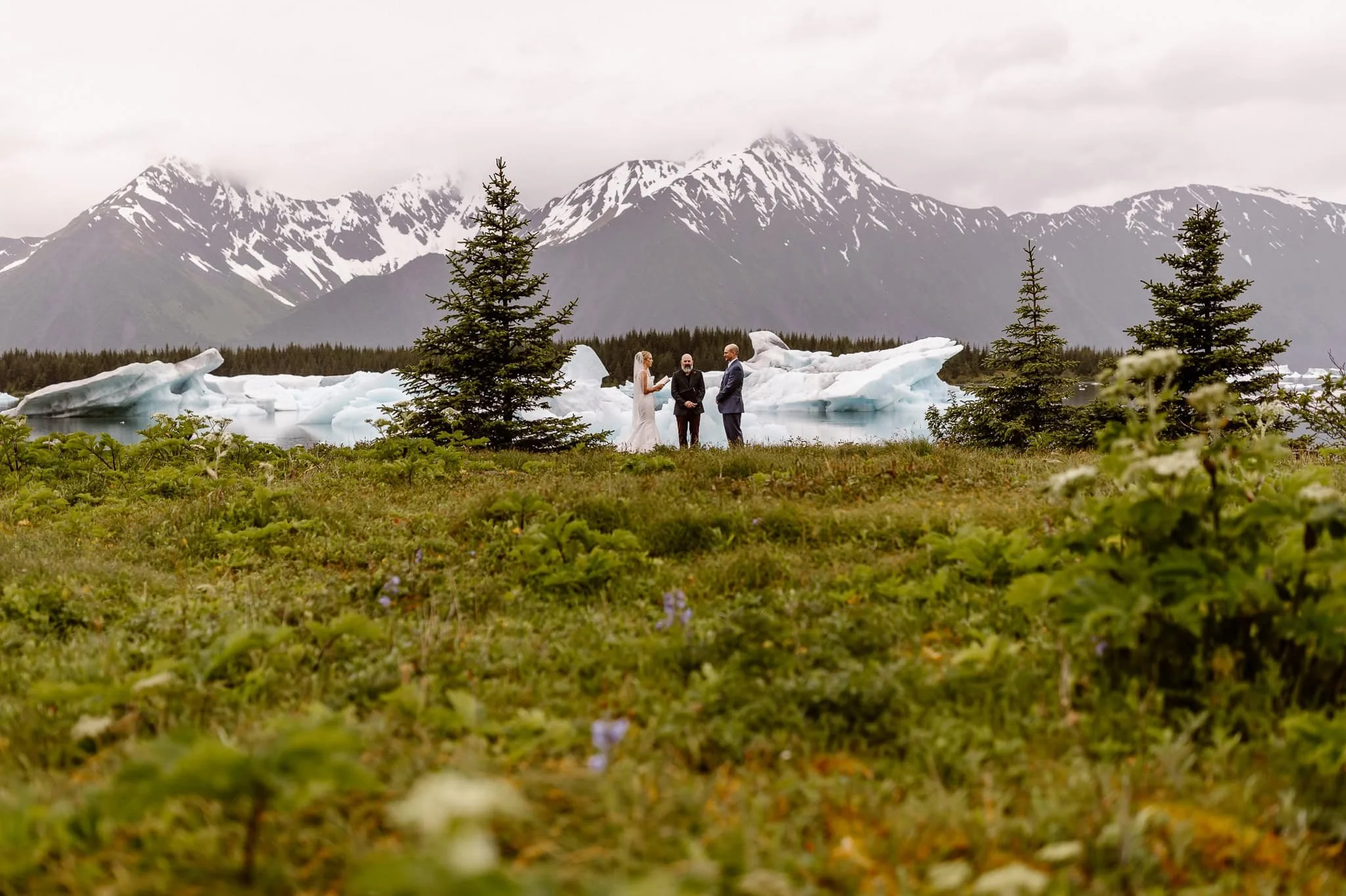 A far-away shot of their ceremony, this image shows the towering mountains in the distance, tops covered in clouds, as well as the light blue icebergs floating in the water below, as the bride and groom recite their private vows on this lush glacier.