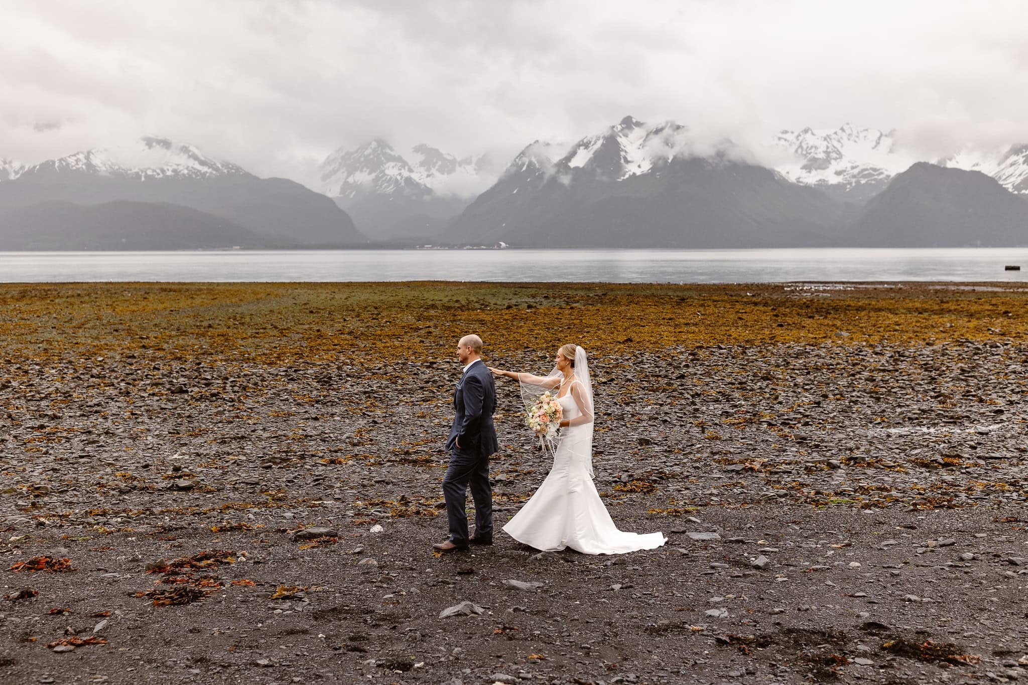 On the shore of Resurrection Bay for their Alaska elopement, the bride stands behind the groom with her bouquet in one hand and the other touching his shoulder. He stands looking away from her in anticipation.