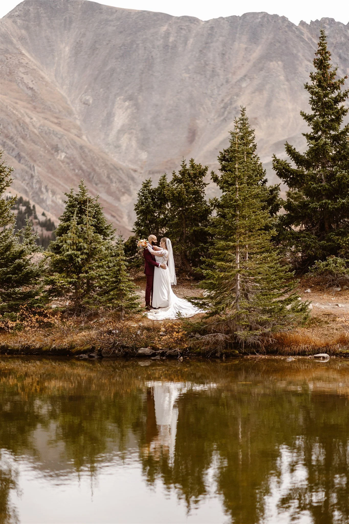 After their ceremony, the bride and groom sneak away for private portraits on the edge of a lake, holding each other closely. Their perfectly romantic reflections appear on the surface of the lake.