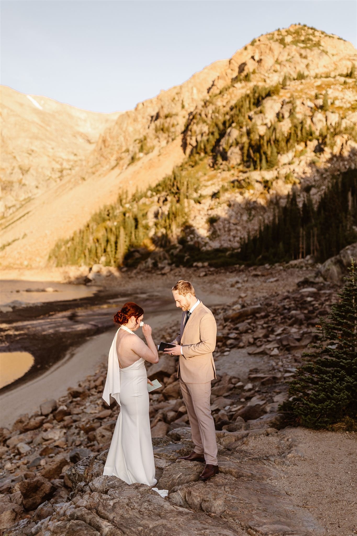 As the sun turns the mountains yellow, the bride wipes tears from her eyes as her groom reads his vows from a leather vow book.