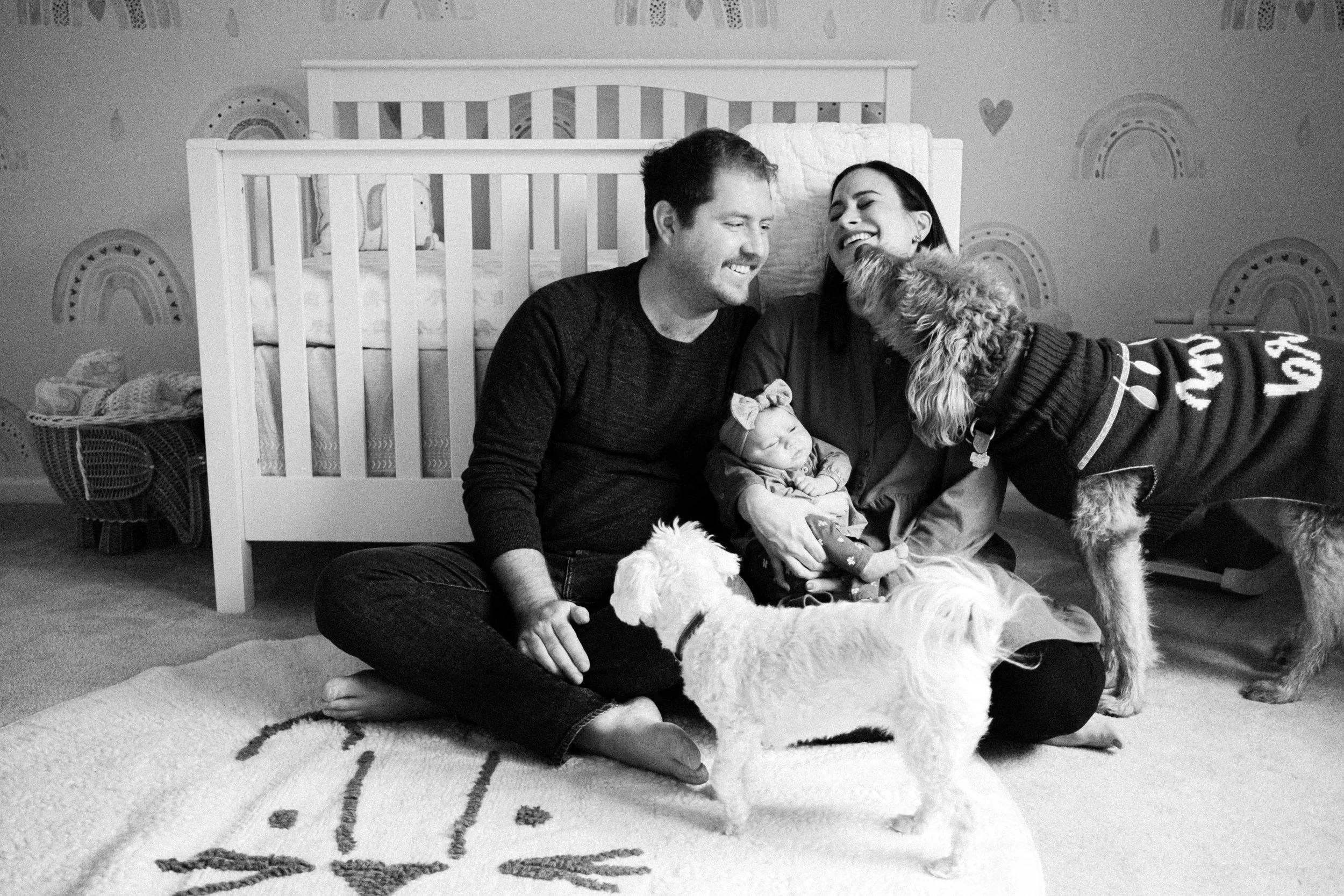 A happy family inside a nursery, sitting on the floor near a crib. A man and woman are smiling at each other, with the woman holding a sleeping baby girl. A small white dog is standing in front of them, looking up at the woman, and a larger dog wearing a sweater is leaning toward the baby.