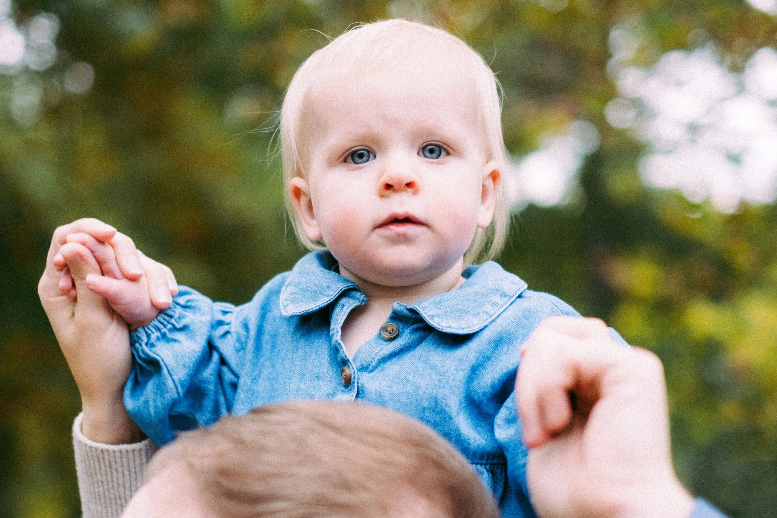 A young child with blonde hair and blue eyes is being held up outdoors, wearing a denim shirt, with a blurred background of trees.