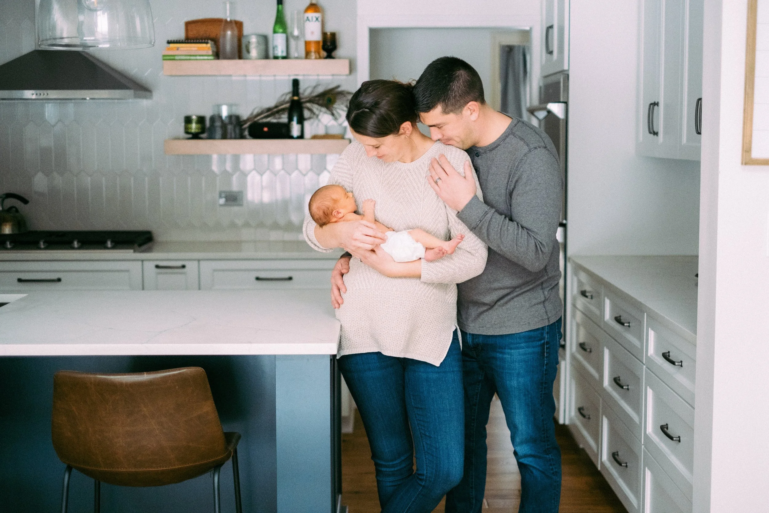 A family family in a modern kitchen, mother holding a newborn baby, father leaning close, all sharing a tender moment.