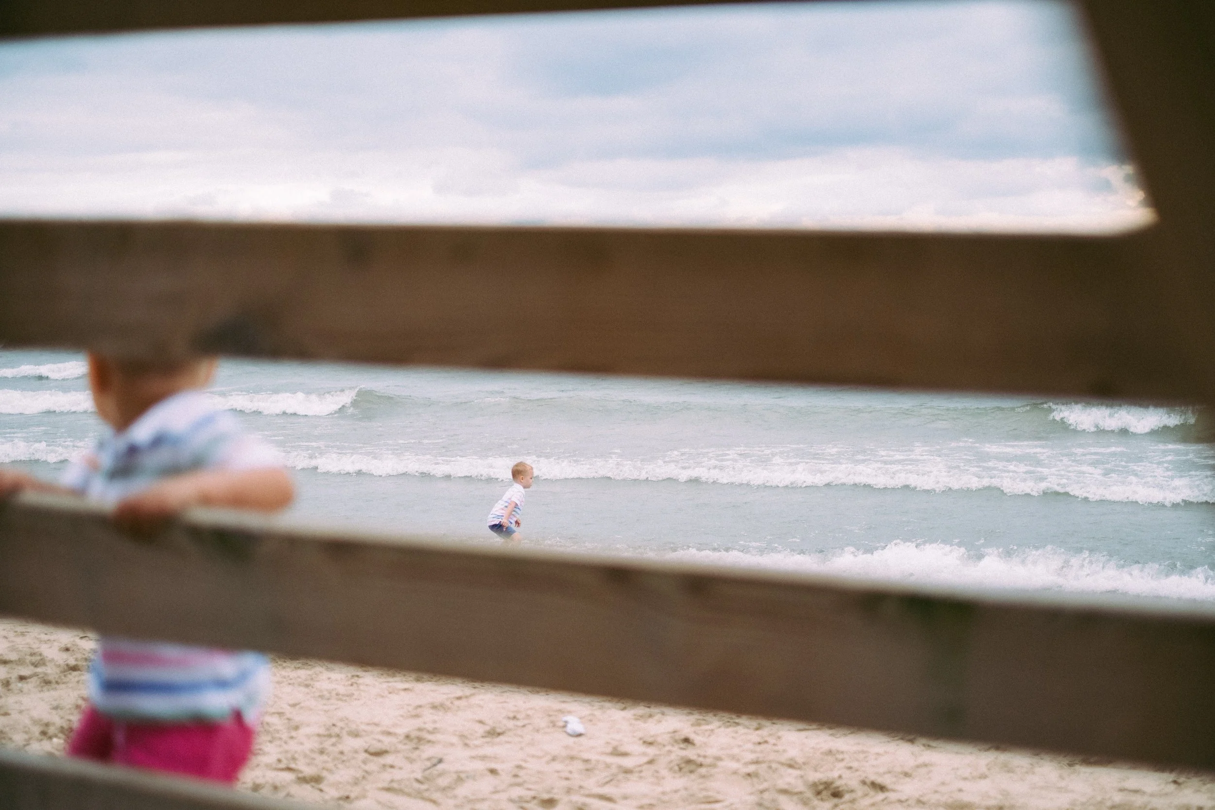 Two young children playing on a beach, seen through wooden slats. The ocean waves and cloudy sky in the background.