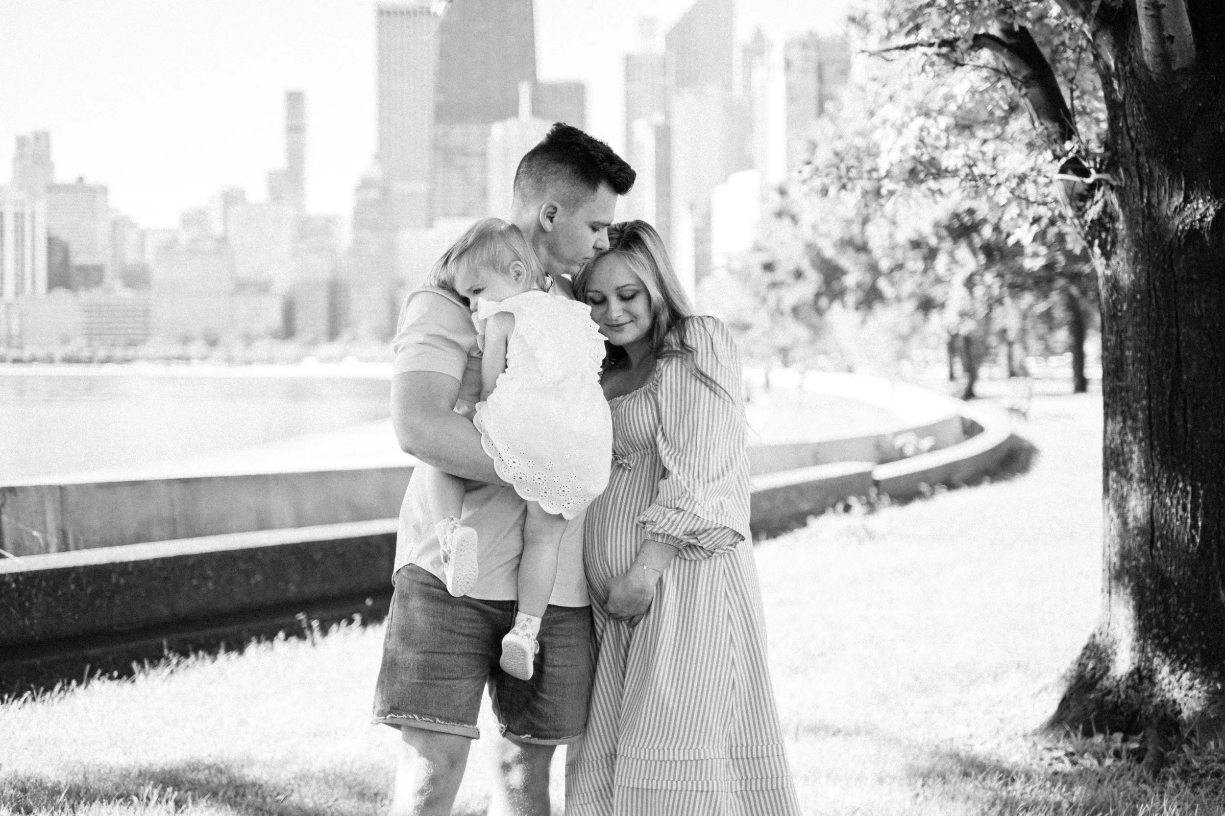 A pregnant woman, a man, and a young girl are standing together outdoors, sharing a tender moment. The man is holding the girl, who is leaning on the pregnant woman's belly. They are standing on a grassy area with city buildings in the background and a large tree nearby.