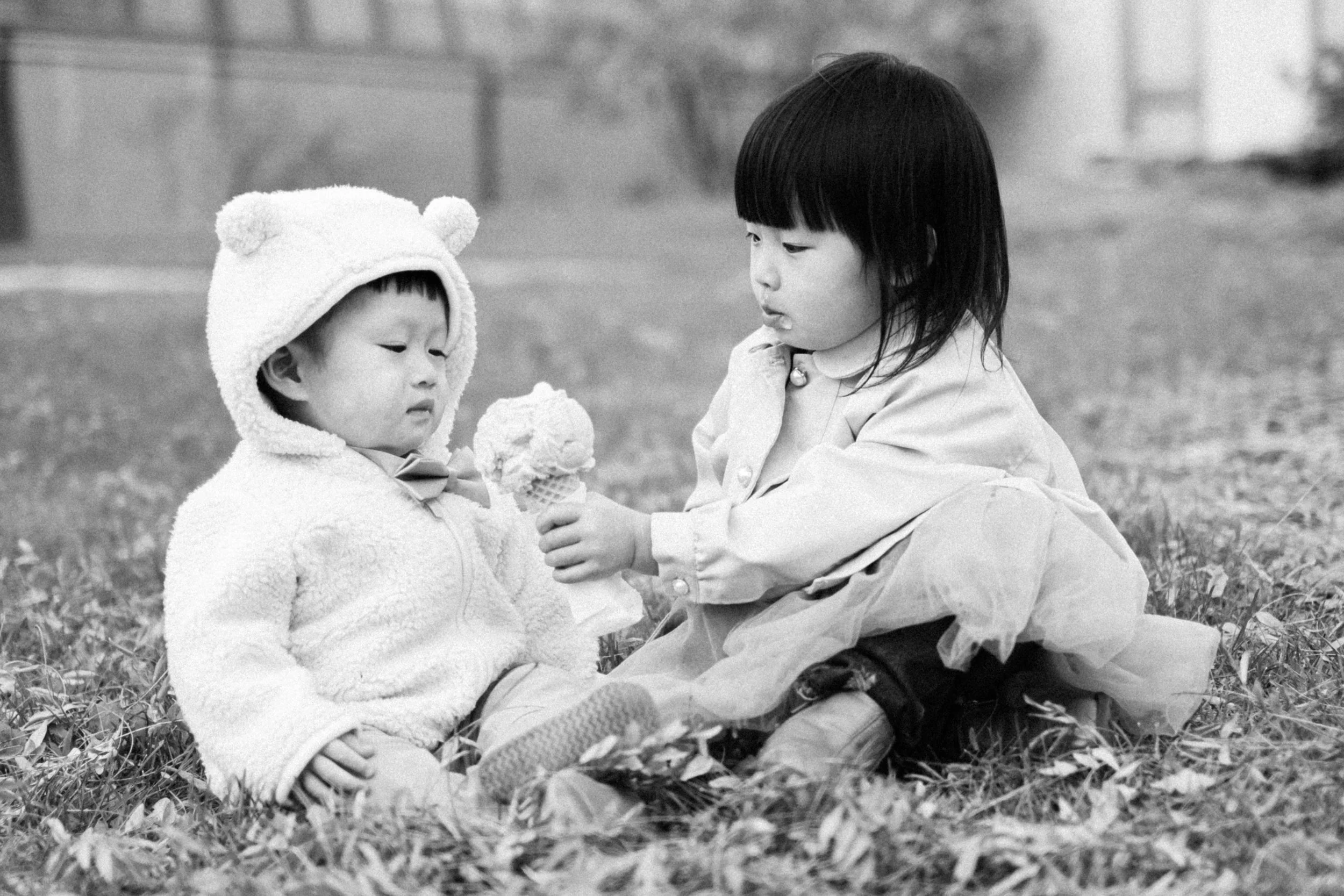 Two young children sit on grass in a park, one offering an ice cream cone to the other. The child on the left wears a fluffy hat with bear ears and a cozy jacket, while the child on the right wears a light-colored jacket and pants.
