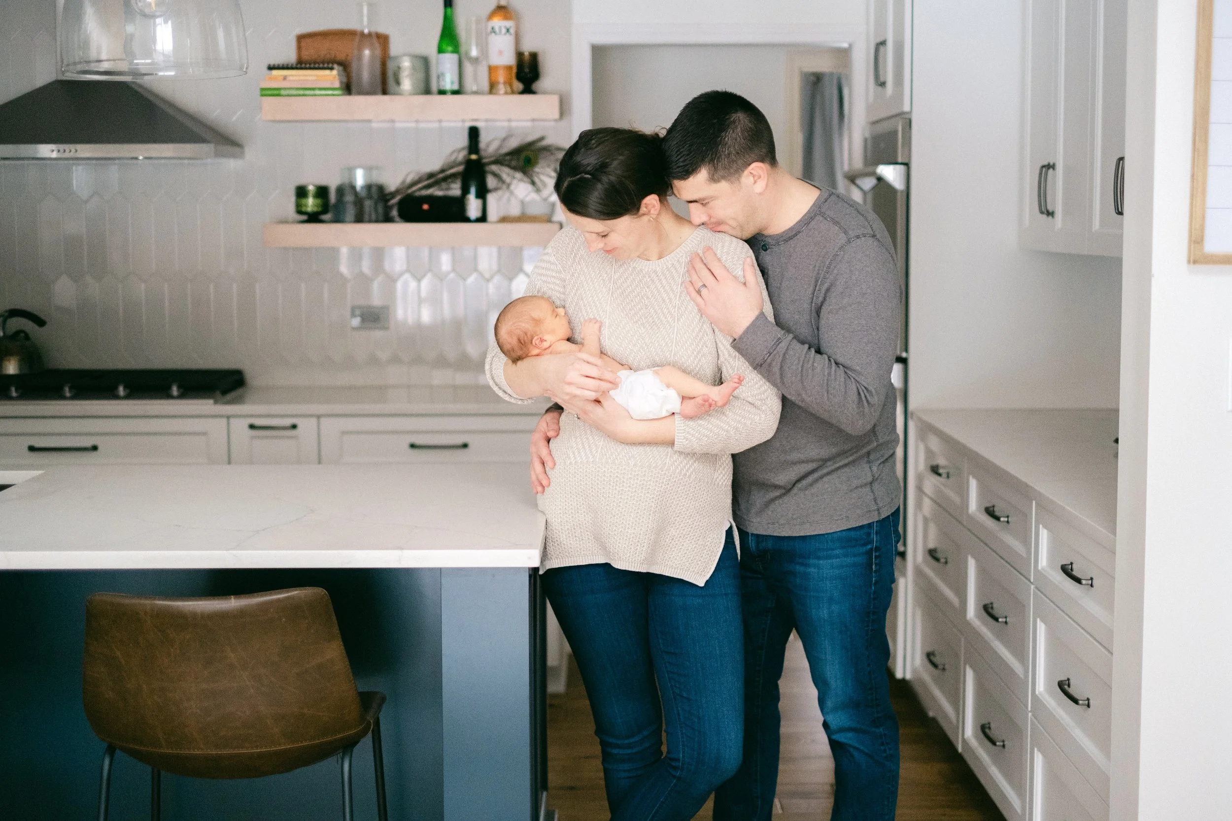 A family family in a modern kitchen, mother holding a newborn baby, father leaning close, all sharing a tender moment.