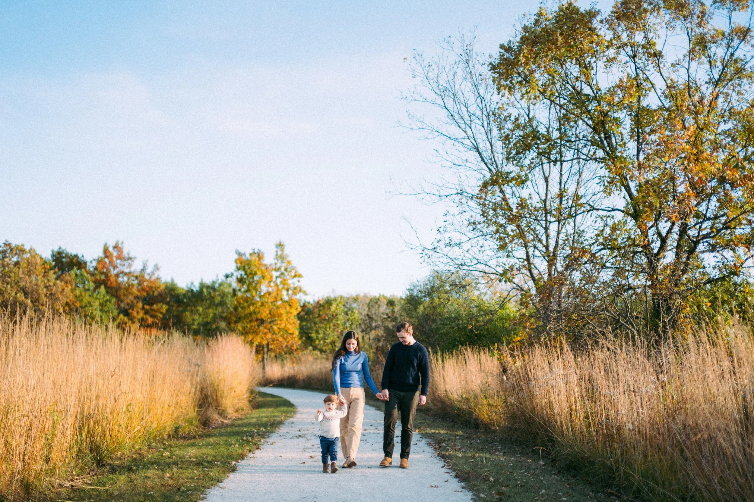 A family of three walking hand in hand on a winding path through a park during autumn, with trees and tall grasses around them.