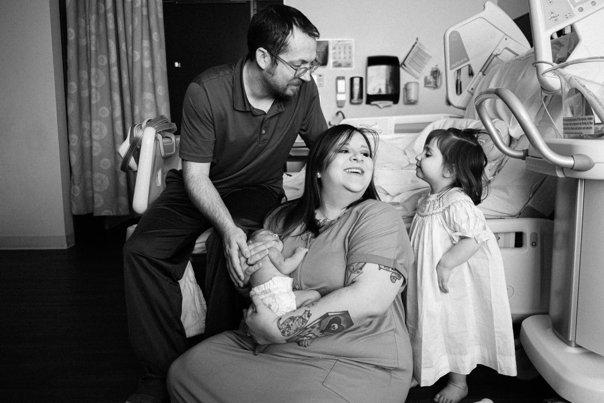 A family in a hospital room, including a mother holding a newborn, a father leaning over, a young girl standing nearby, all smiling and interacting.