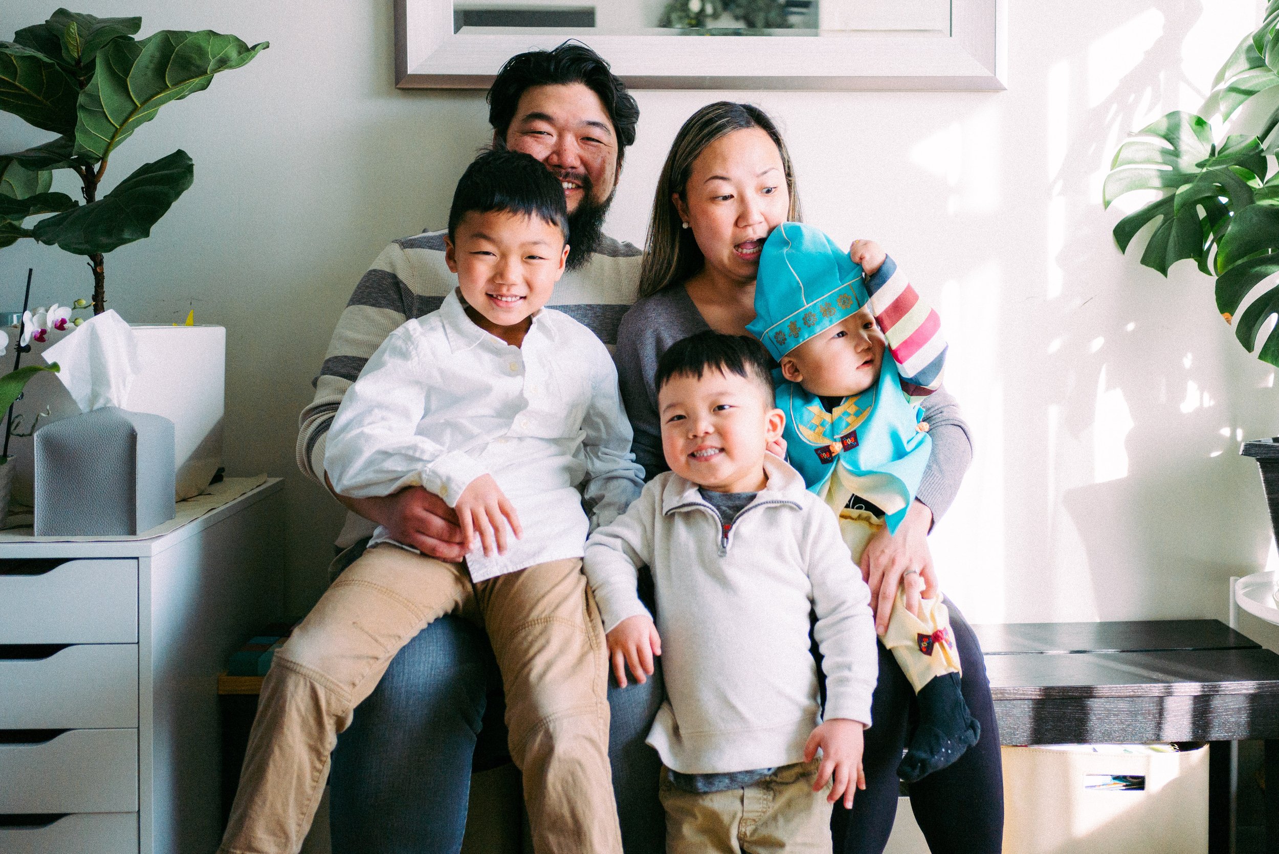 A family of five with two adults and three children sitting together in a living room, smiling and enjoying each other's company.