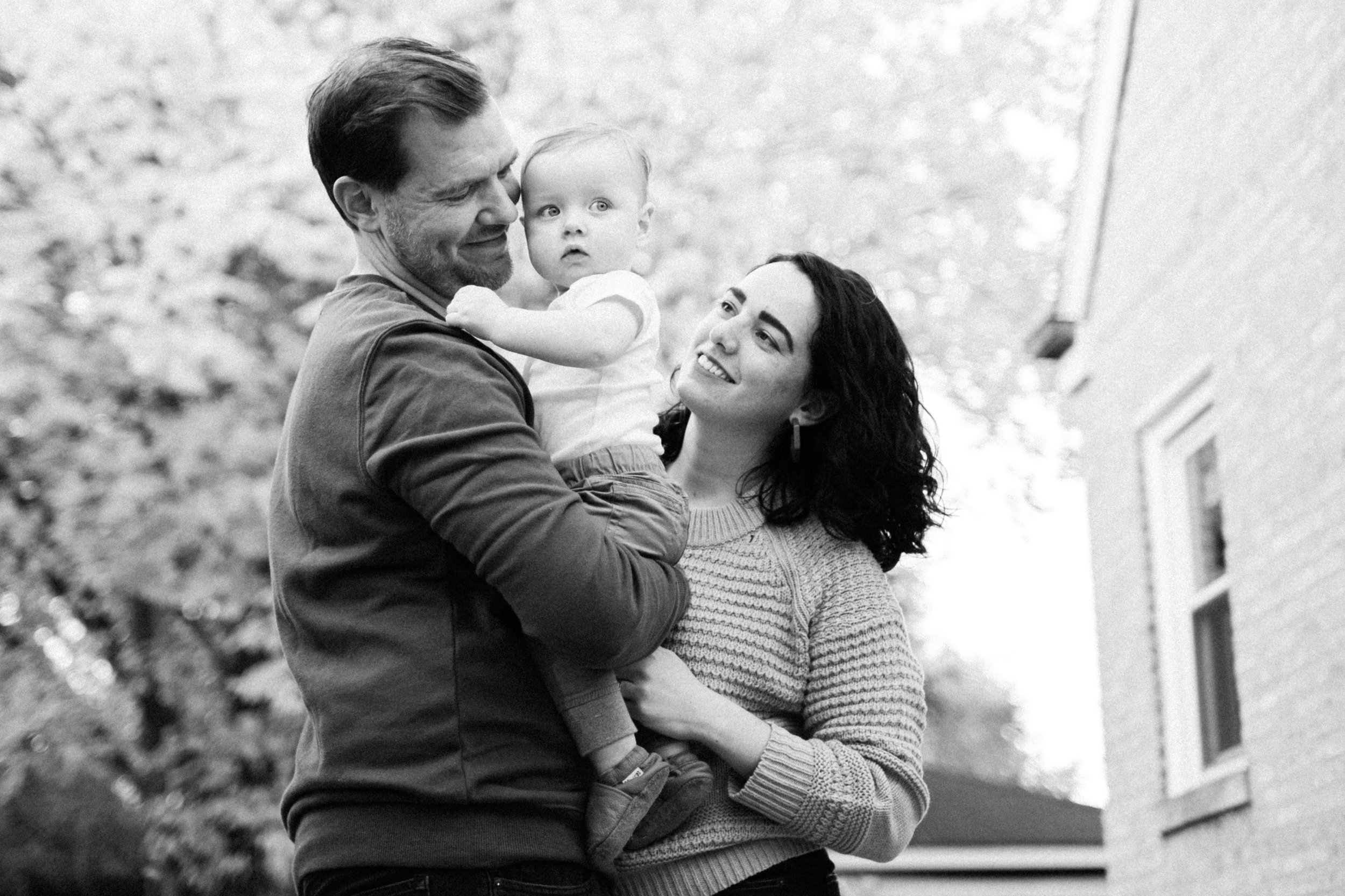 A family of three outside, with a man holding a young child and a woman looking at the child, all smiling.