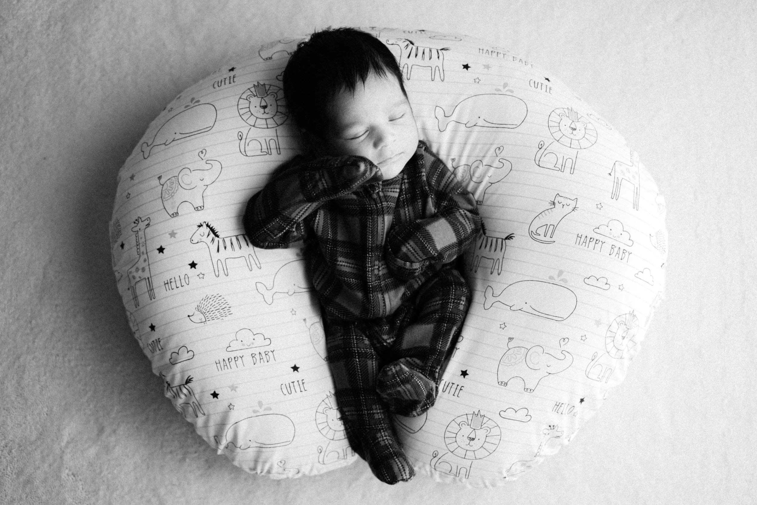 A sleeping baby lying on a round, cushioned pillow with animal illustrations and words like 'hello', 'happy baby', and 'cutie' in black and white.