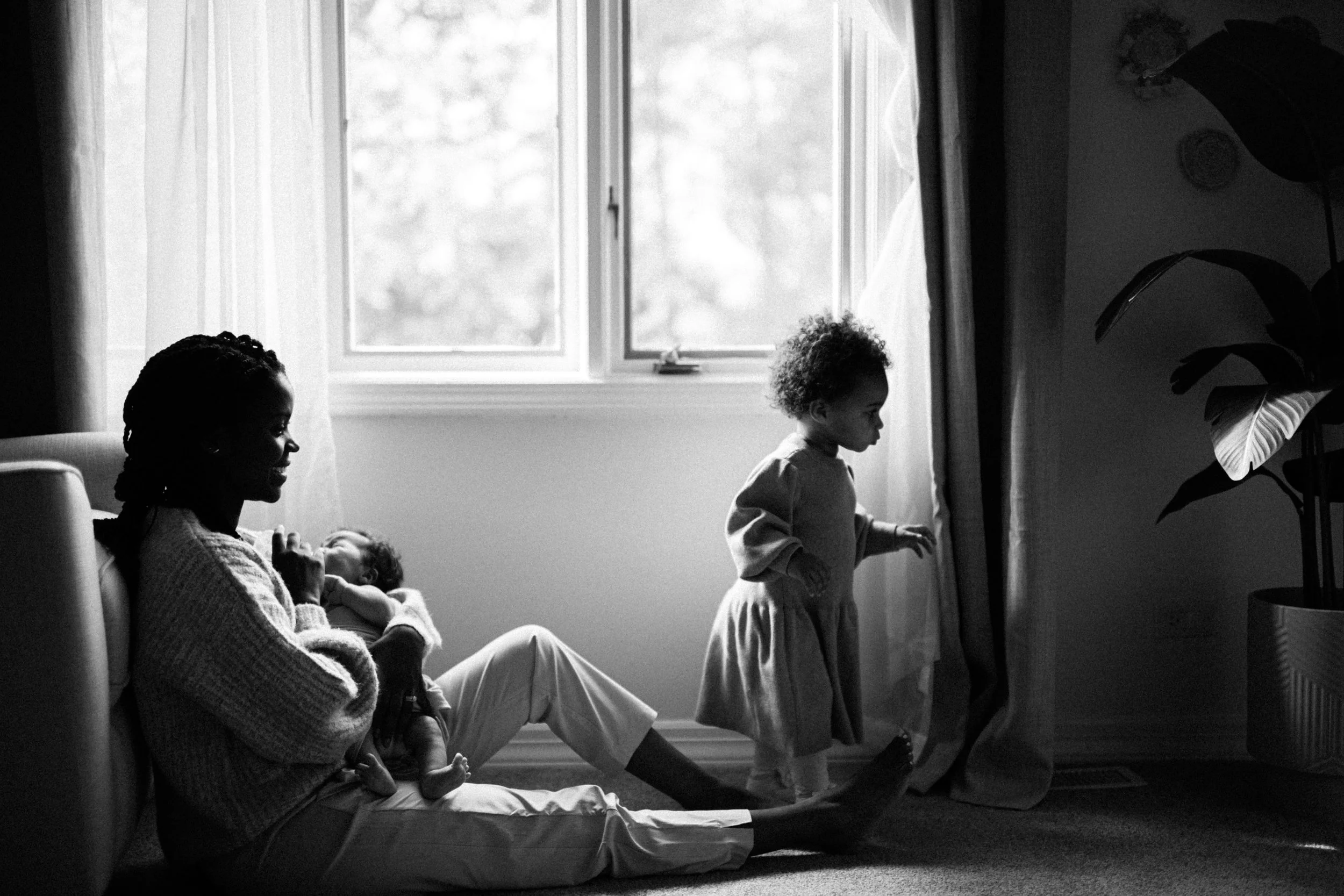 A woman with two children in a room, with one child sitting on her lap and the other standing near a window, looking outside. The image is in black and white.