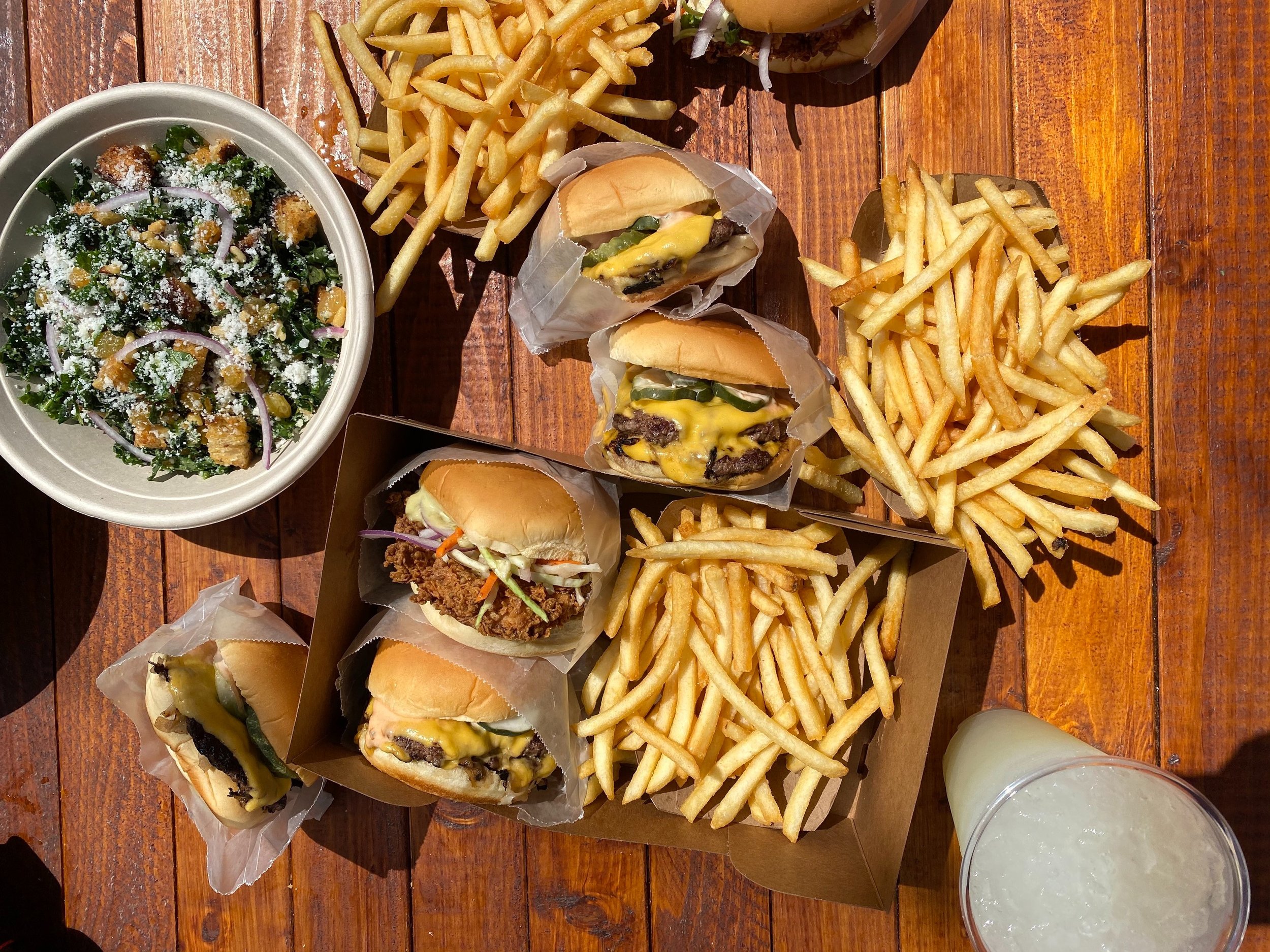  wooden tabletop view of food including burgers, fried chicken sandwiches, french fries and a kale salad 