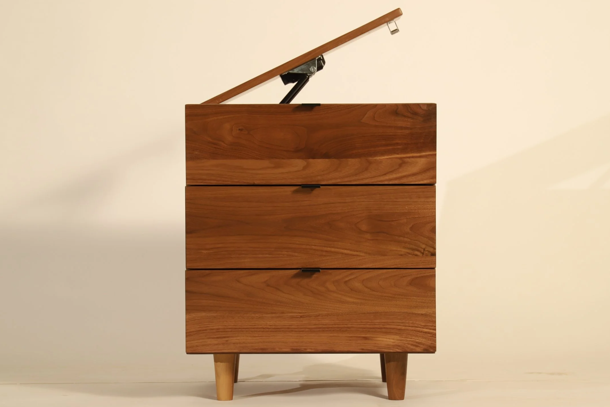 A wooden dresser with four drawers and a drop-down top, with the top partially open and supported by a hydraulic hinge, against a plain light-colored wall.