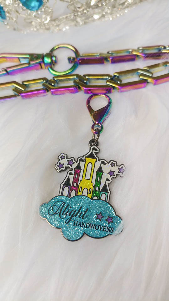 Photo ID: a fuzzy white background with a crown at the top, a chain below followed by a charm of the alight Handwovens castle