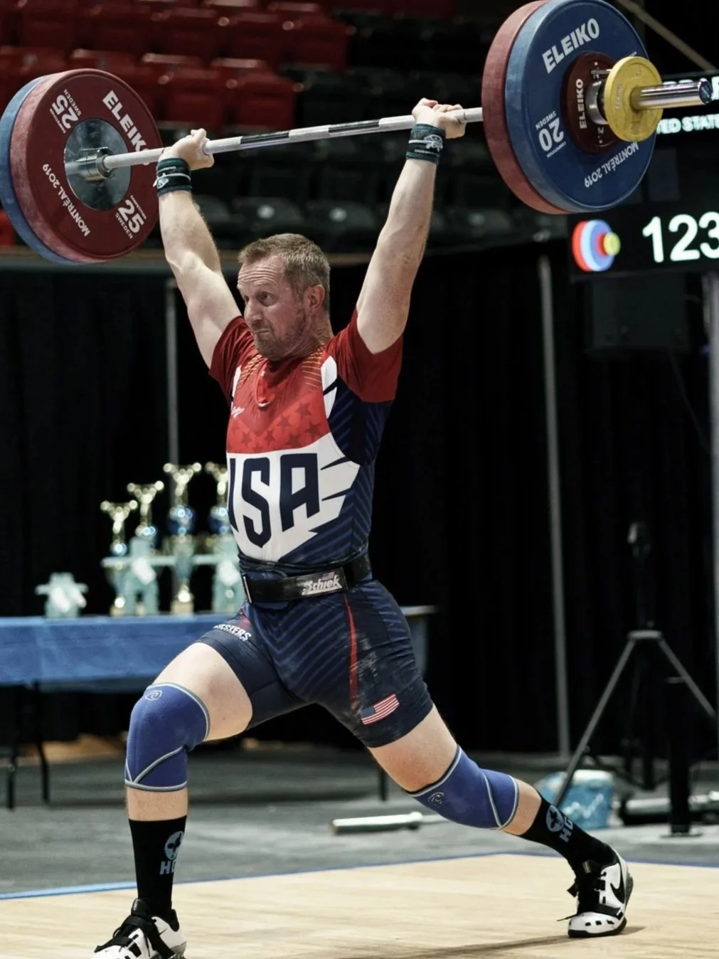 A male athlete in USA team uniform lifting a loaded barbell overhead during a weightlifting competition.