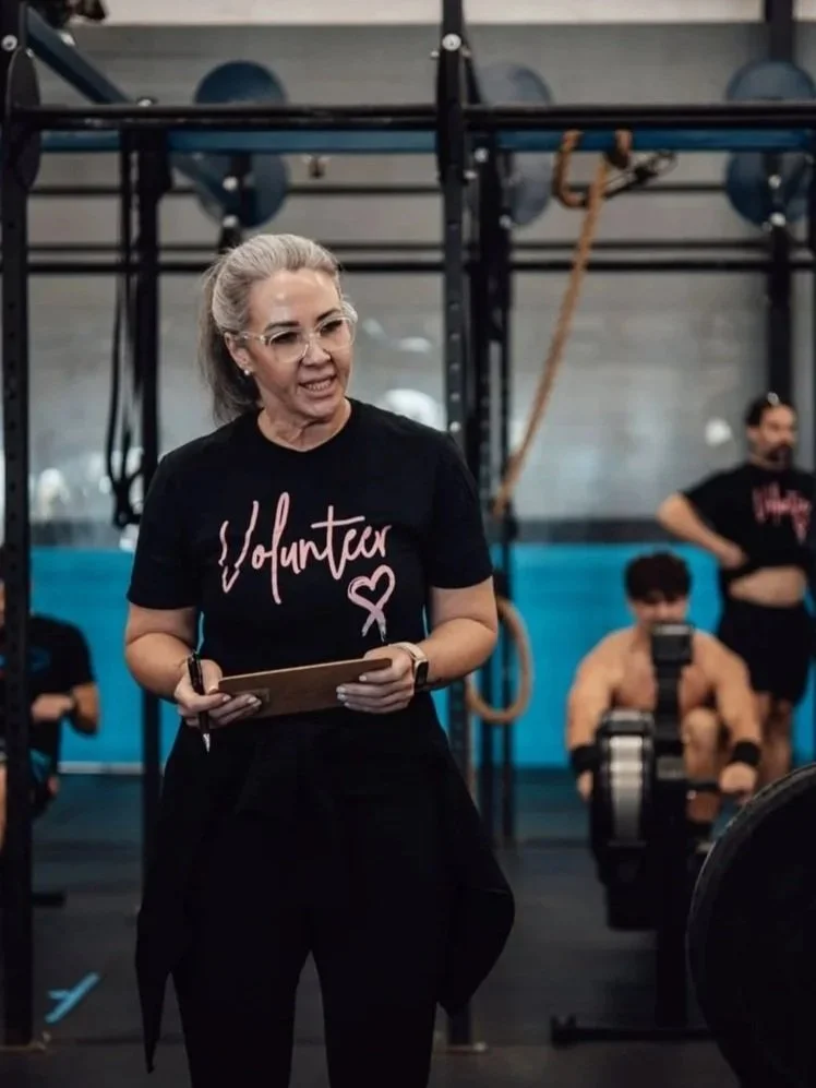 A woman with gray hair and glasses, wearing a black volunteer T-shirt, stands in a gym holding a clipboard. Behind her, people are working out, including a shirtless man on rowing machines and others engaging in fitness activities.