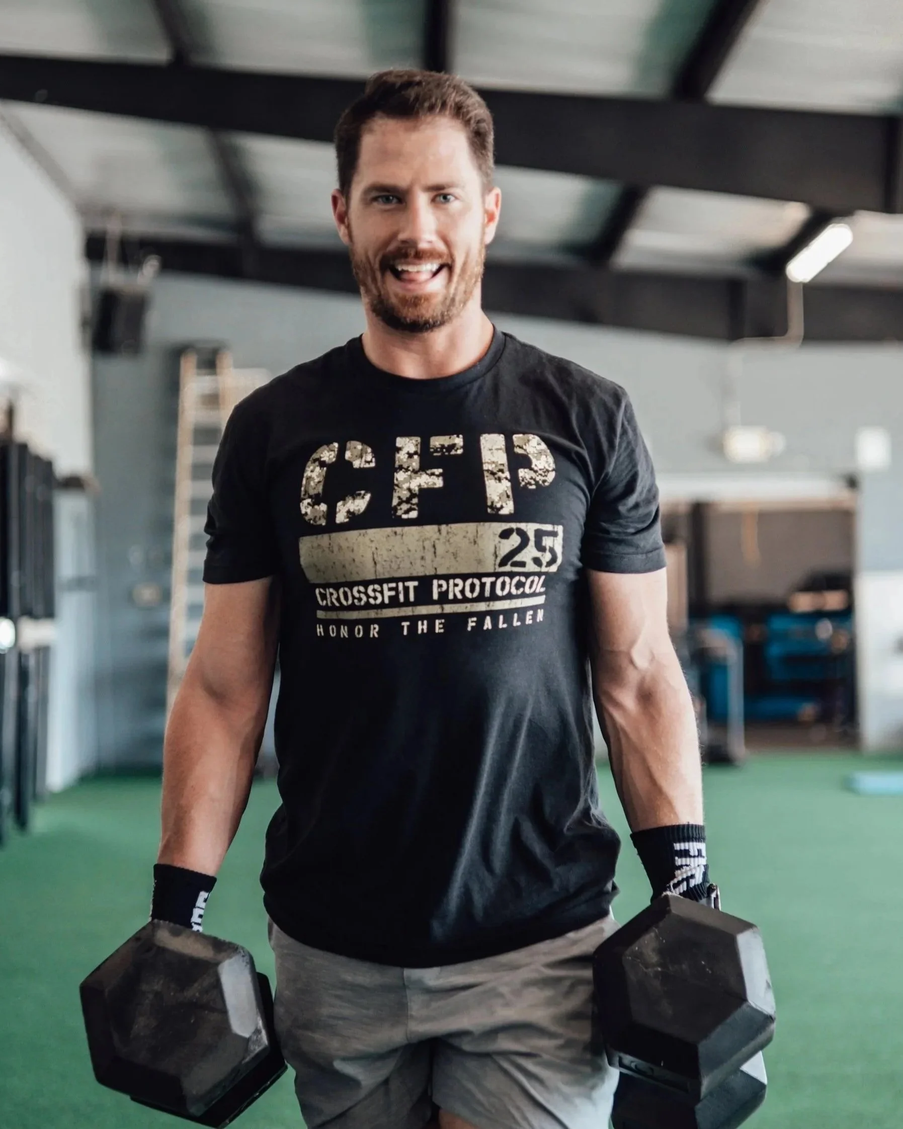 A man with short brown hair and a beard, smiling and wearing a black T-shirt with CrossFit branding, holding a pair of black dumbbells in a gym.
