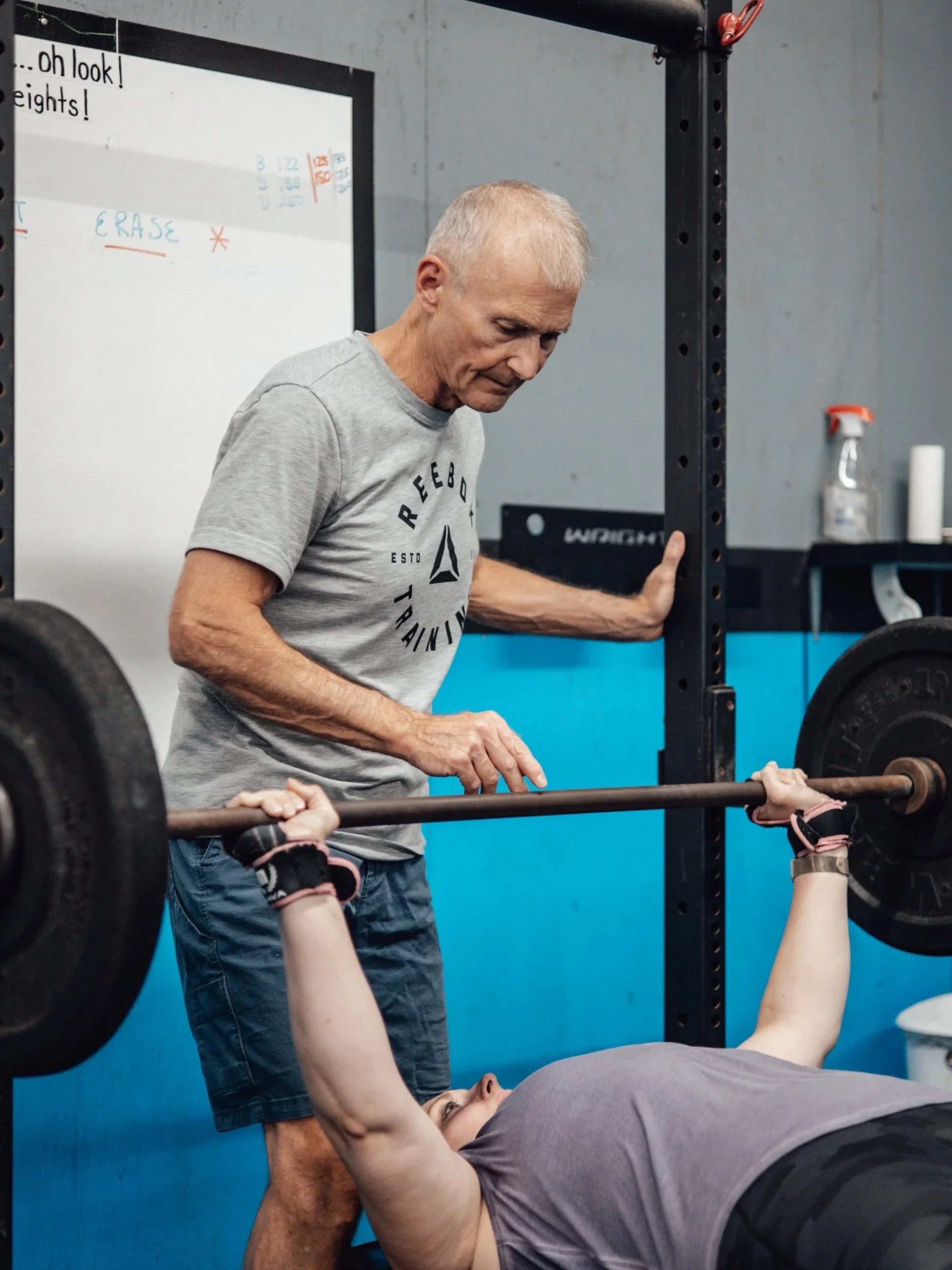 Older man spotting younger woman lifting a barbell during workout in a gym.
